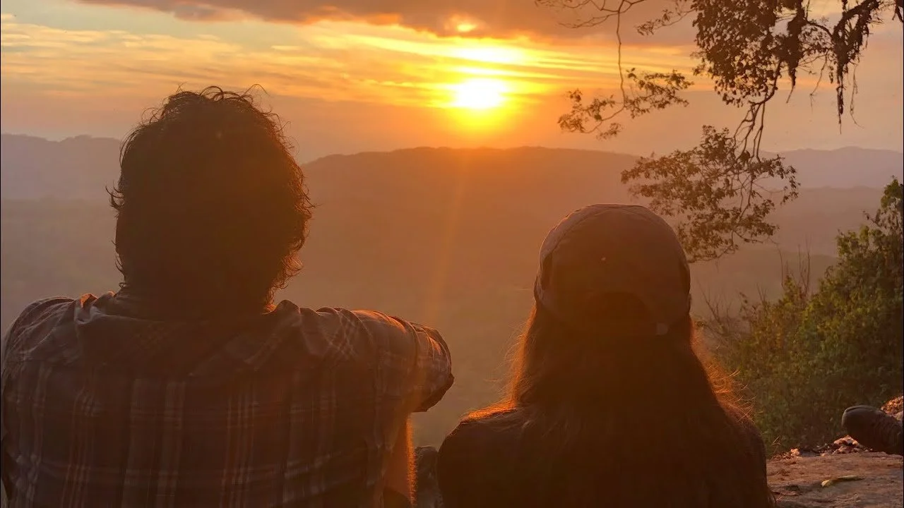 Two people sitting outdoors watching a sunset over mountains and trees.