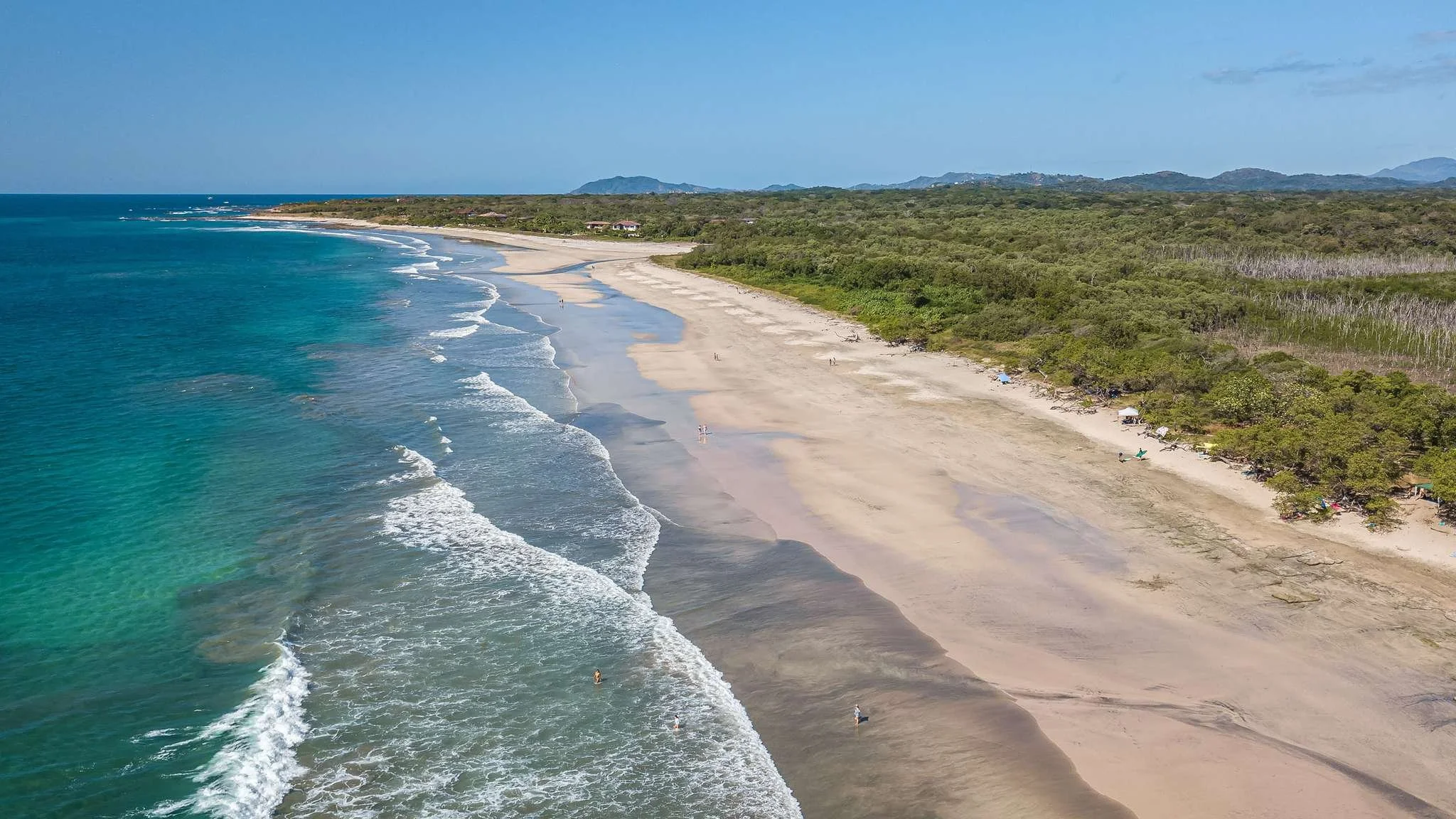 Aerial view of a long, sandy beach with turquoise ocean waves and lush green forest in the background.