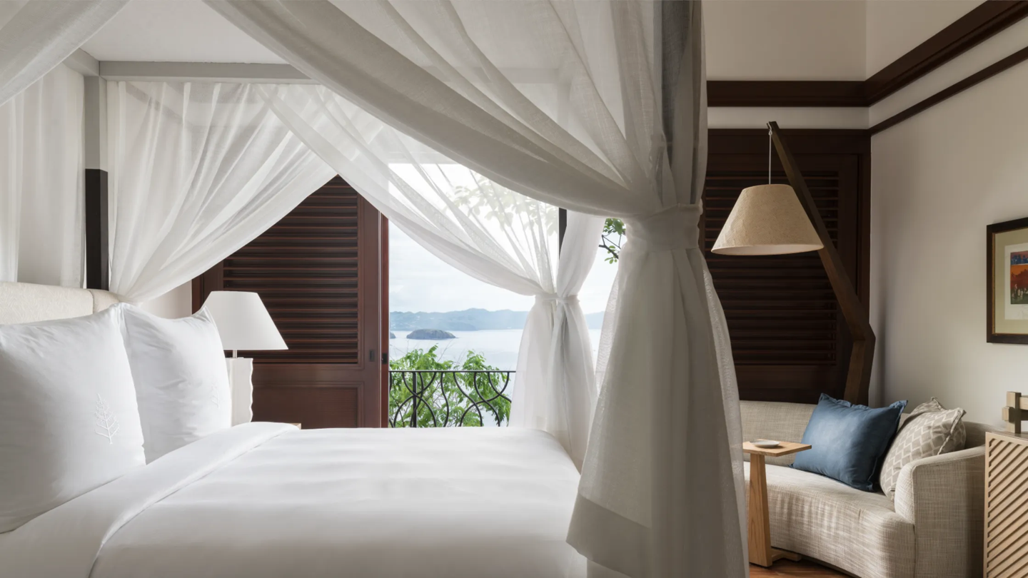A luxurious bedroom with a four-poster bed draped with white curtains, overlooking a balcony with a water and mountain view, with a side sitting area featuring a sofa, pillows, and a lamp.
