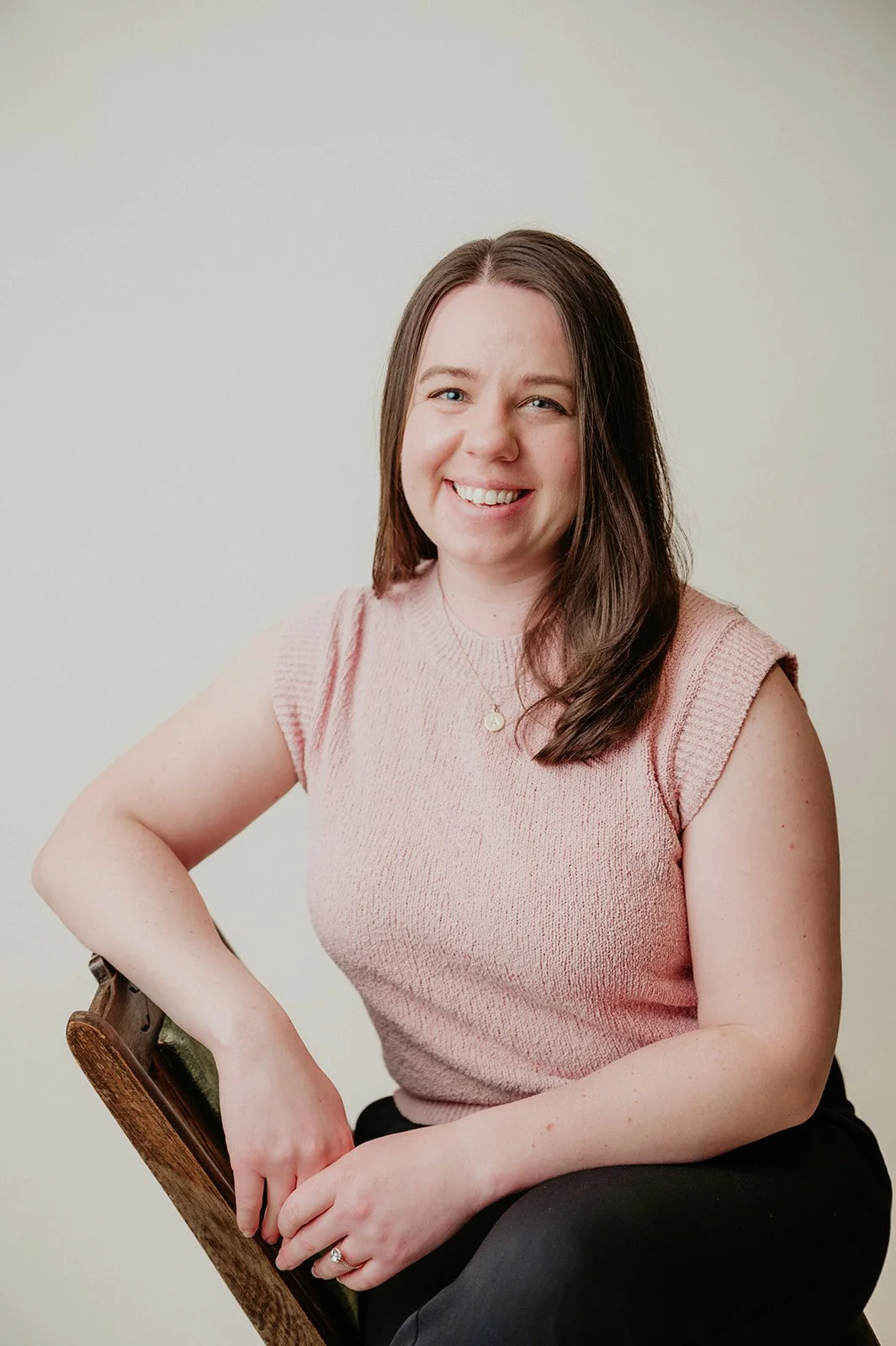 Ali Thiel, A woman with brown hair smiling while sitting on a chair against a plain, light-colored background.