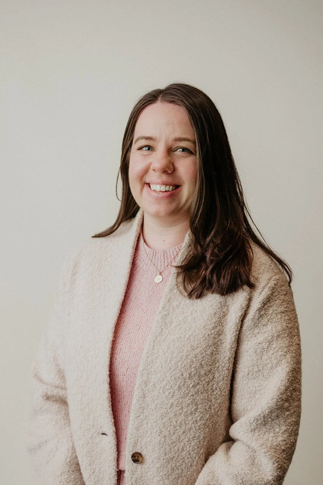 Ali Thiel, A young woman with long brown hair, smiling, wearing a light pink sweater and a cream-colored coat, standing against a plain off-white wall.