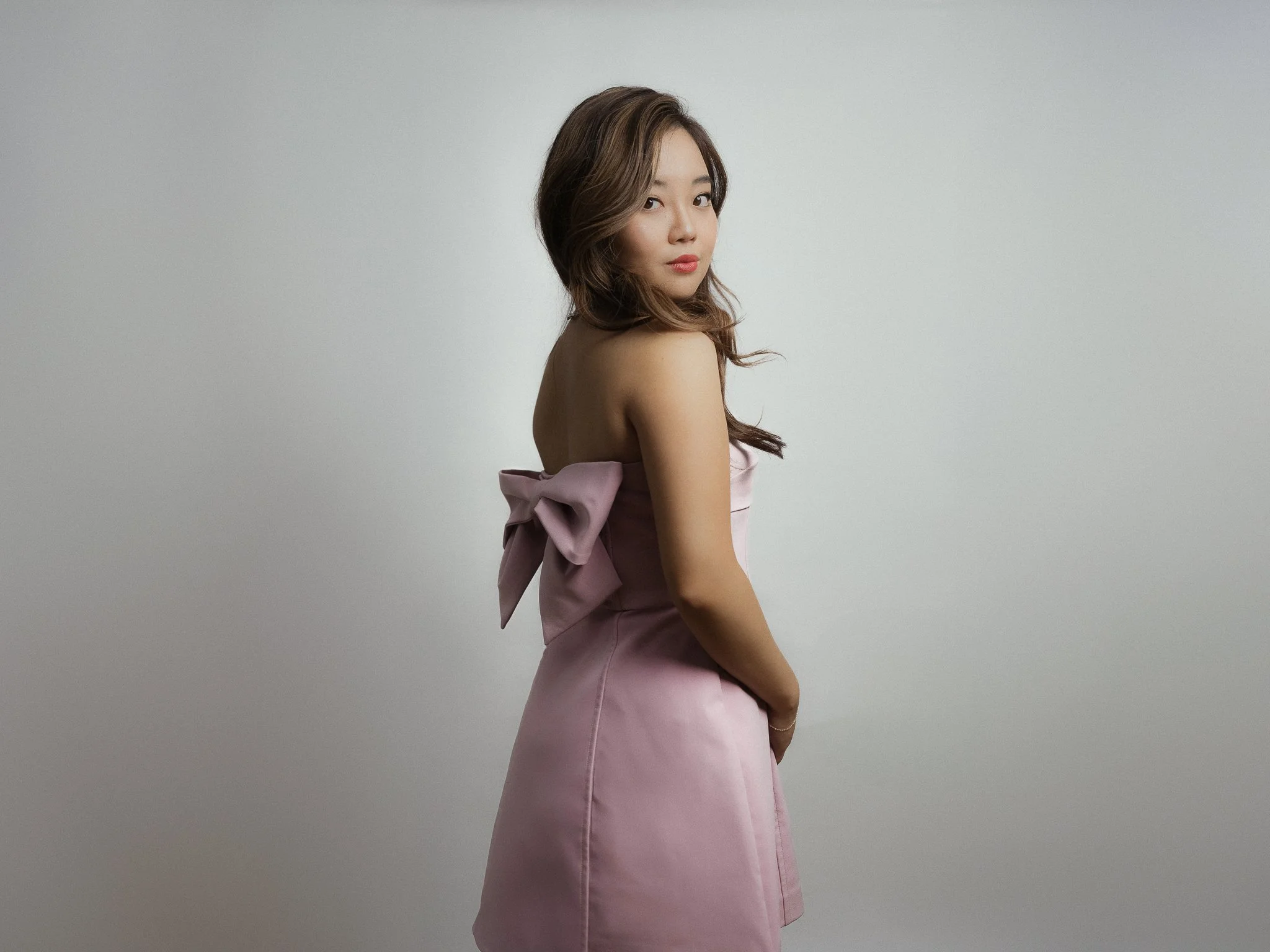 A young woman with long wavy hair wearing a strapless pink dress with a large bow on the back, standing against a plain gray wall, looking over her shoulder.