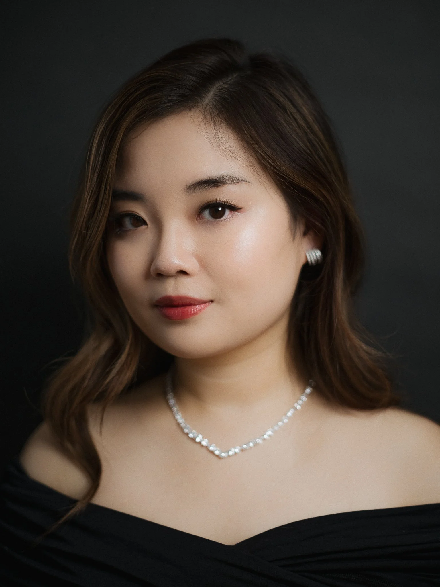 Portrait of a woman with shoulder-length brown hair, wearing a black off-shoulder top, pearl earrings, and a diamond necklace, against a dark background.