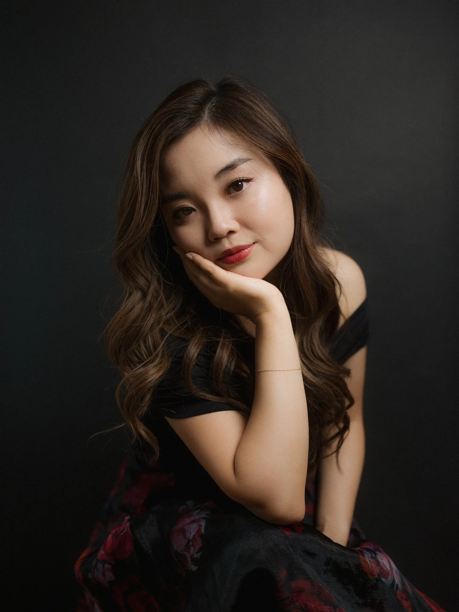 Portrait of a young woman with long wavy brown hair, lightly smiling, resting her head on her hand against a dark background.