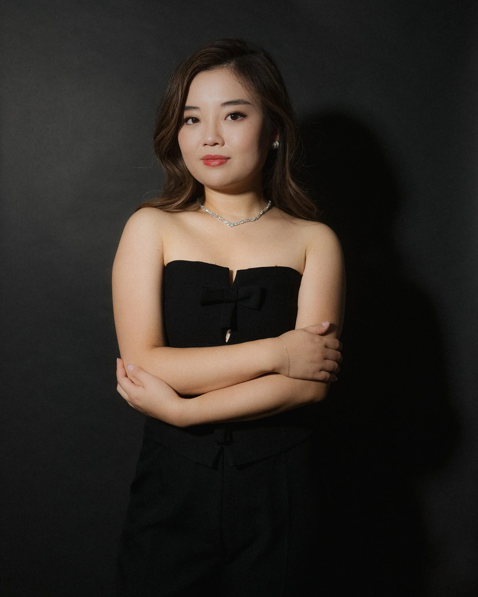 A woman with shoulder-length brown hair in a strapless black dress with a decorative bow, standing against a dark background, with arms crossed and a confident expression.