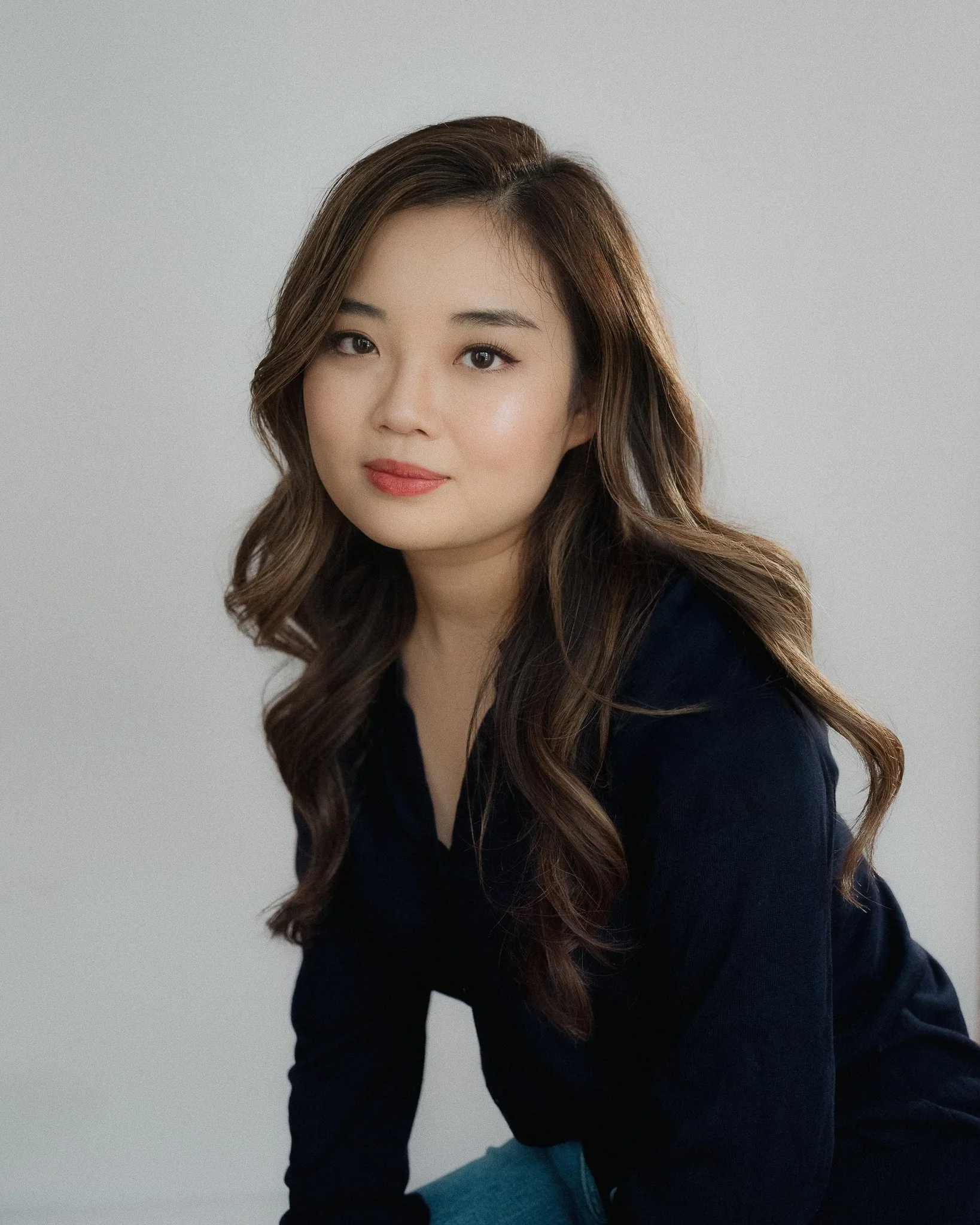 Portrait of a young woman with wavy brown hair, wearing a navy blue top, posing against a plain gray background.