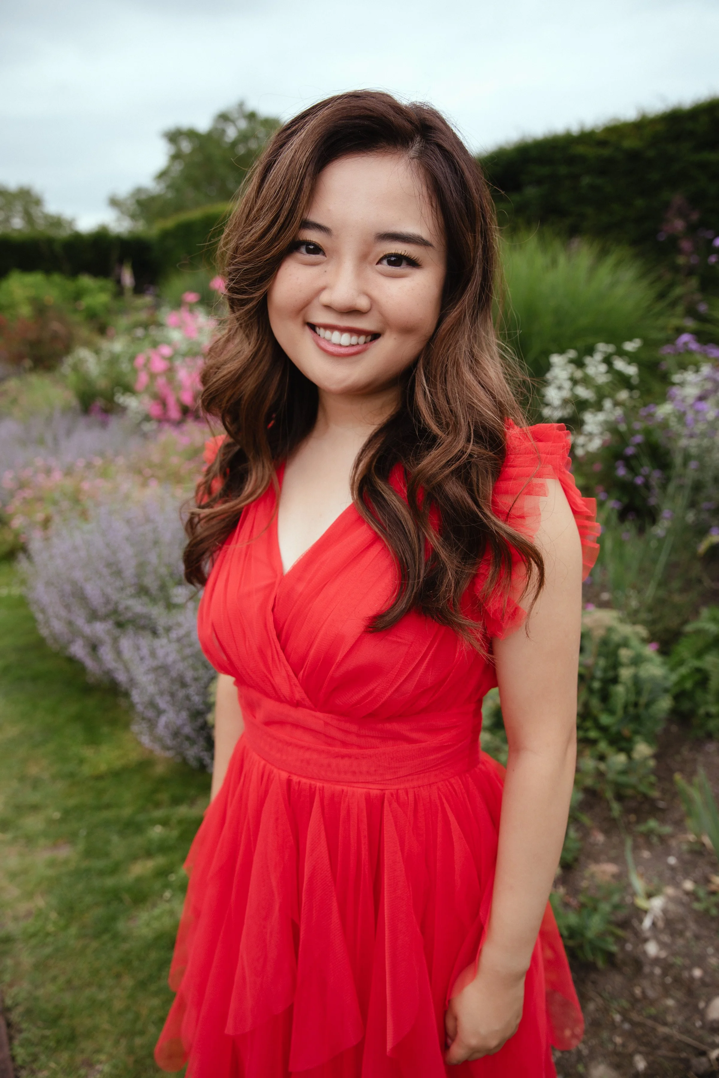 A woman with long, wavy brown hair smiling outdoors in a red dress in a garden with pink, purple, and white flowers.