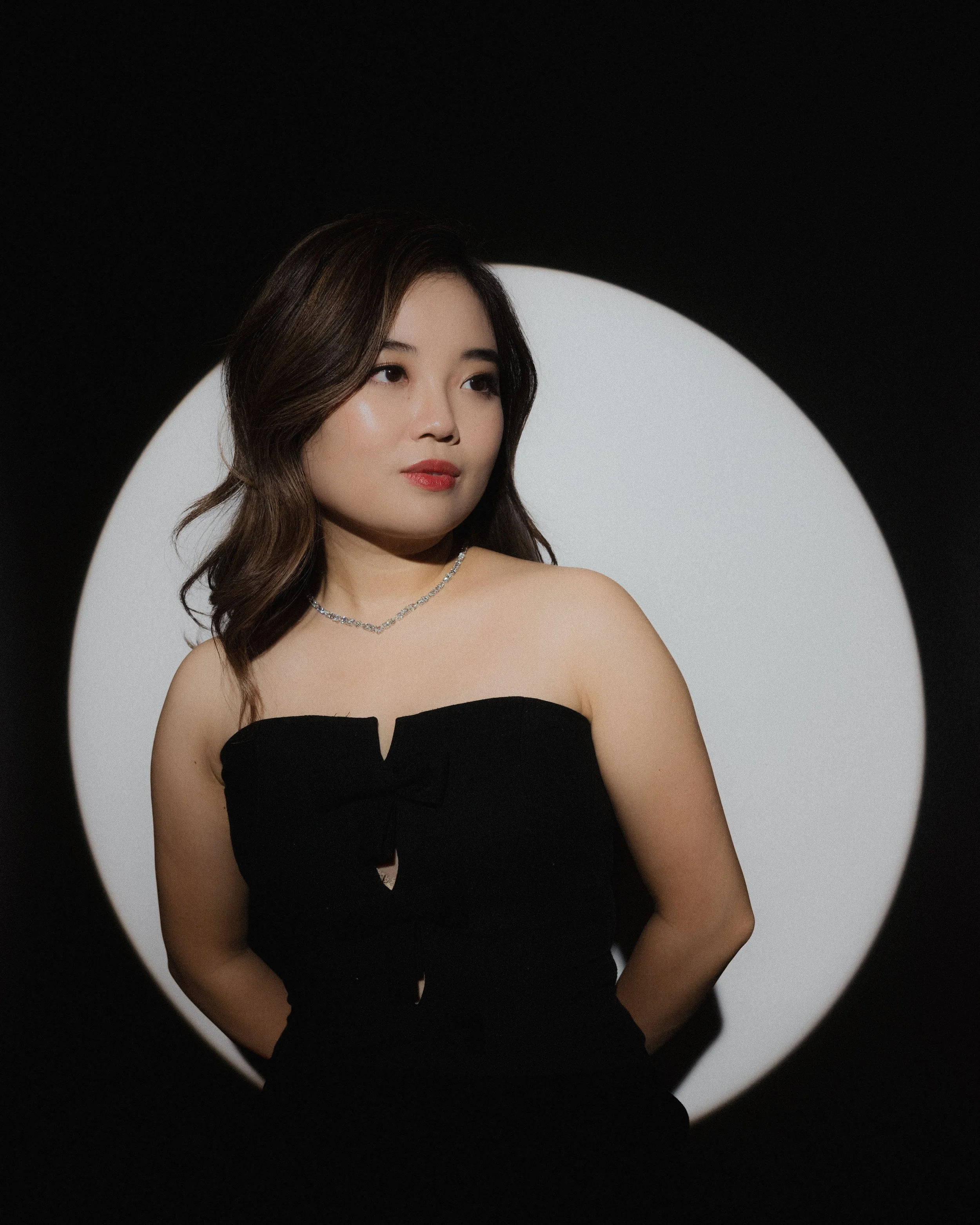 A woman with shoulder-length brown hair, wearing a strapless black dress and a silver necklace, stands in front of a circular white background with a black border.