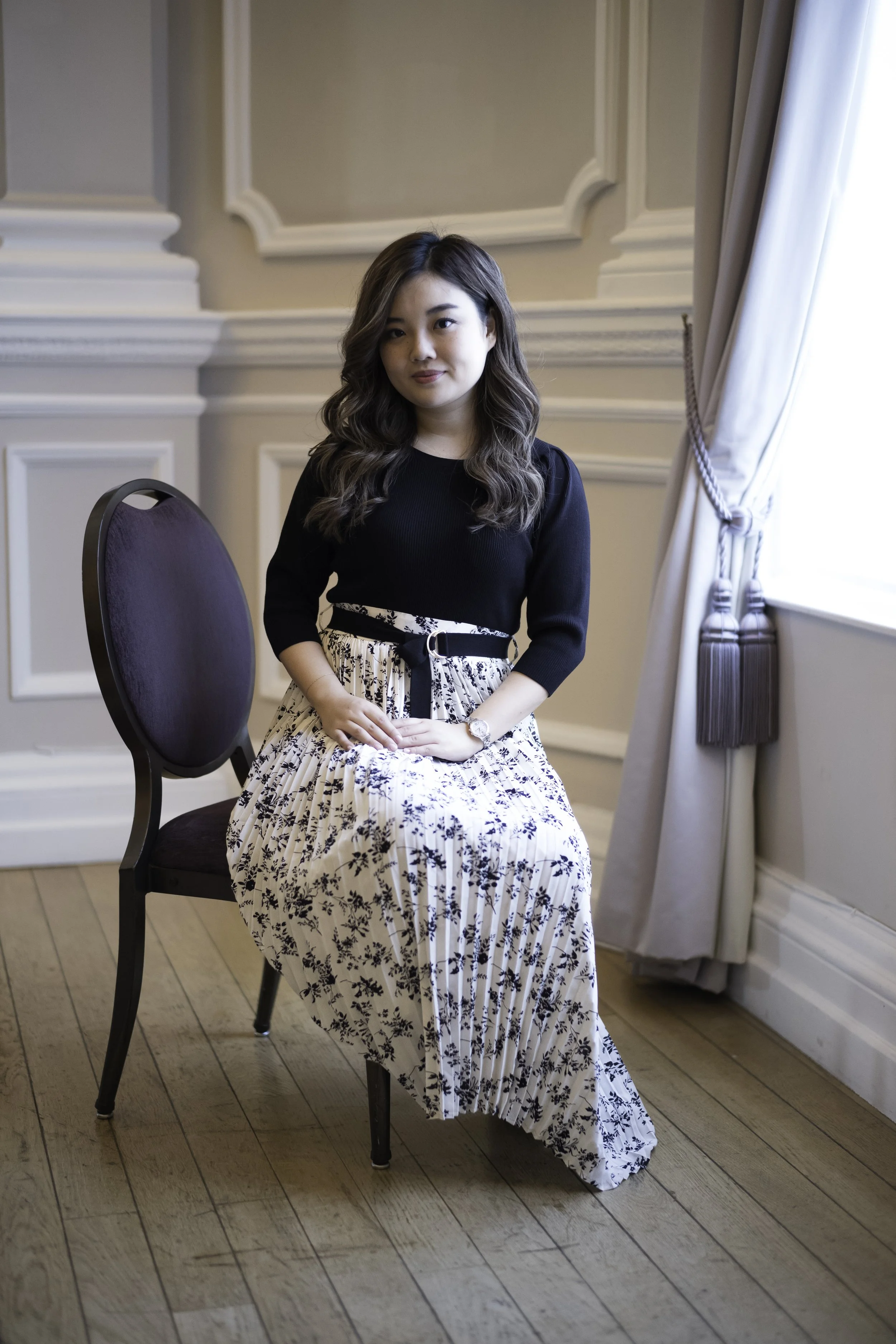 A woman with long wavy hair wearing a black top and a long white skirt with black floral print, sitting on a dark purple chair in a well-lit room with beige walls and a window with curtains.