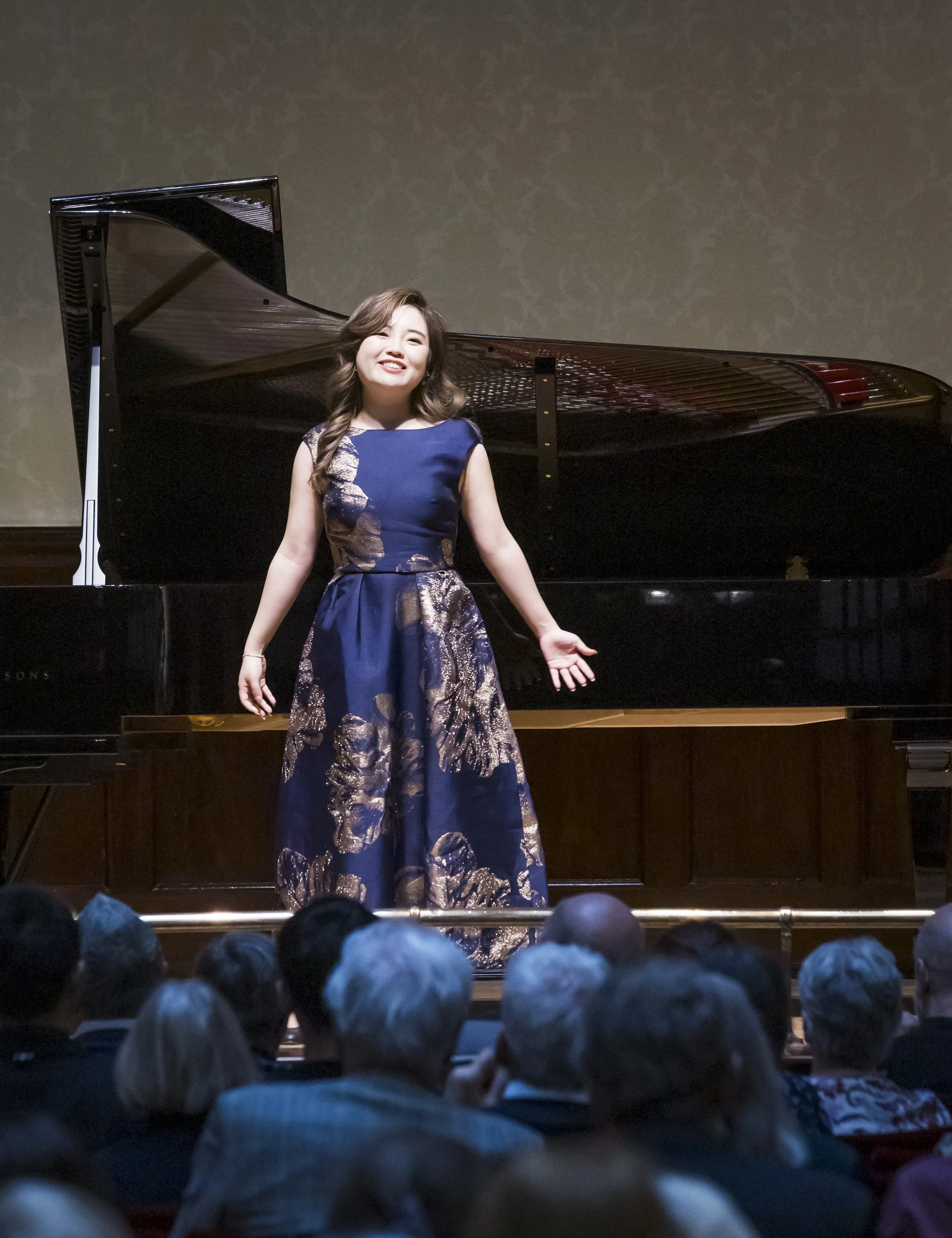 A woman in a elegant navy blue and gold dress stands on stage in front of a grand piano, performing in front of an audience.