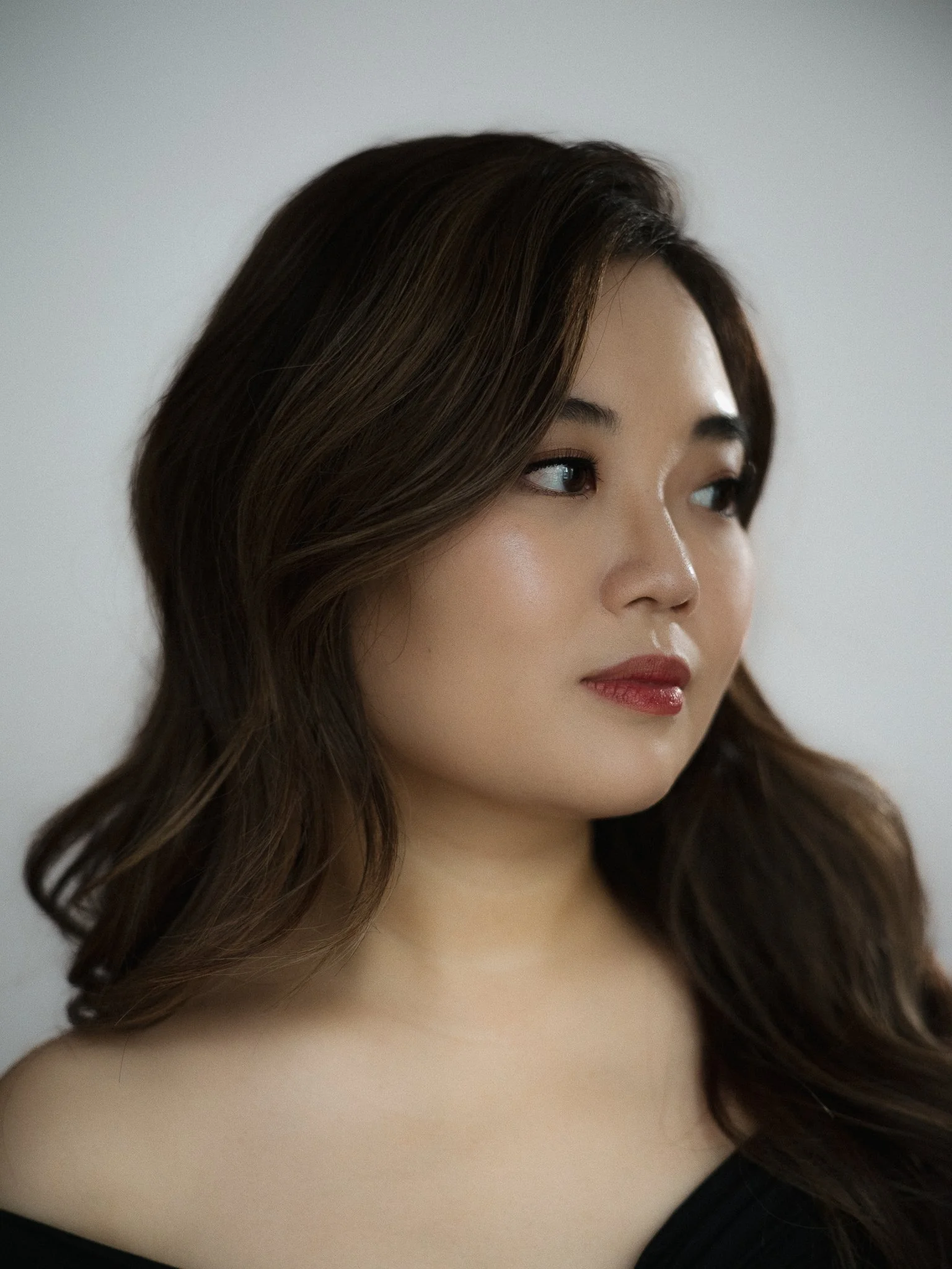 Close-up of a young woman with long wavy brown hair, wearing light makeup with red lipstick, looking to her left against a plain white background.