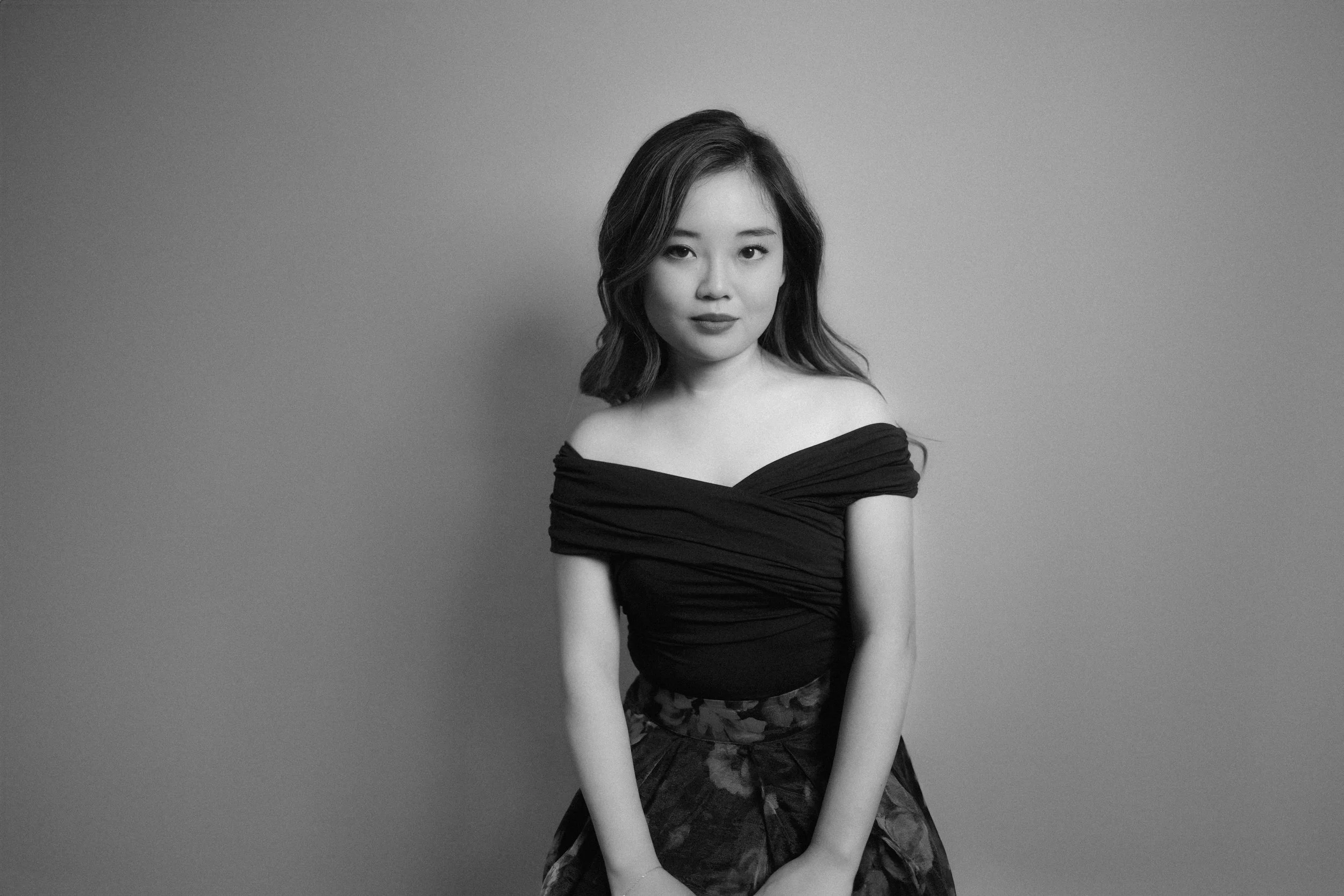 A black and white photo of a young woman with shoulder-length wavy hair, wearing an off-the-shoulder black top and a patterned skirt, standing against a plain wall.