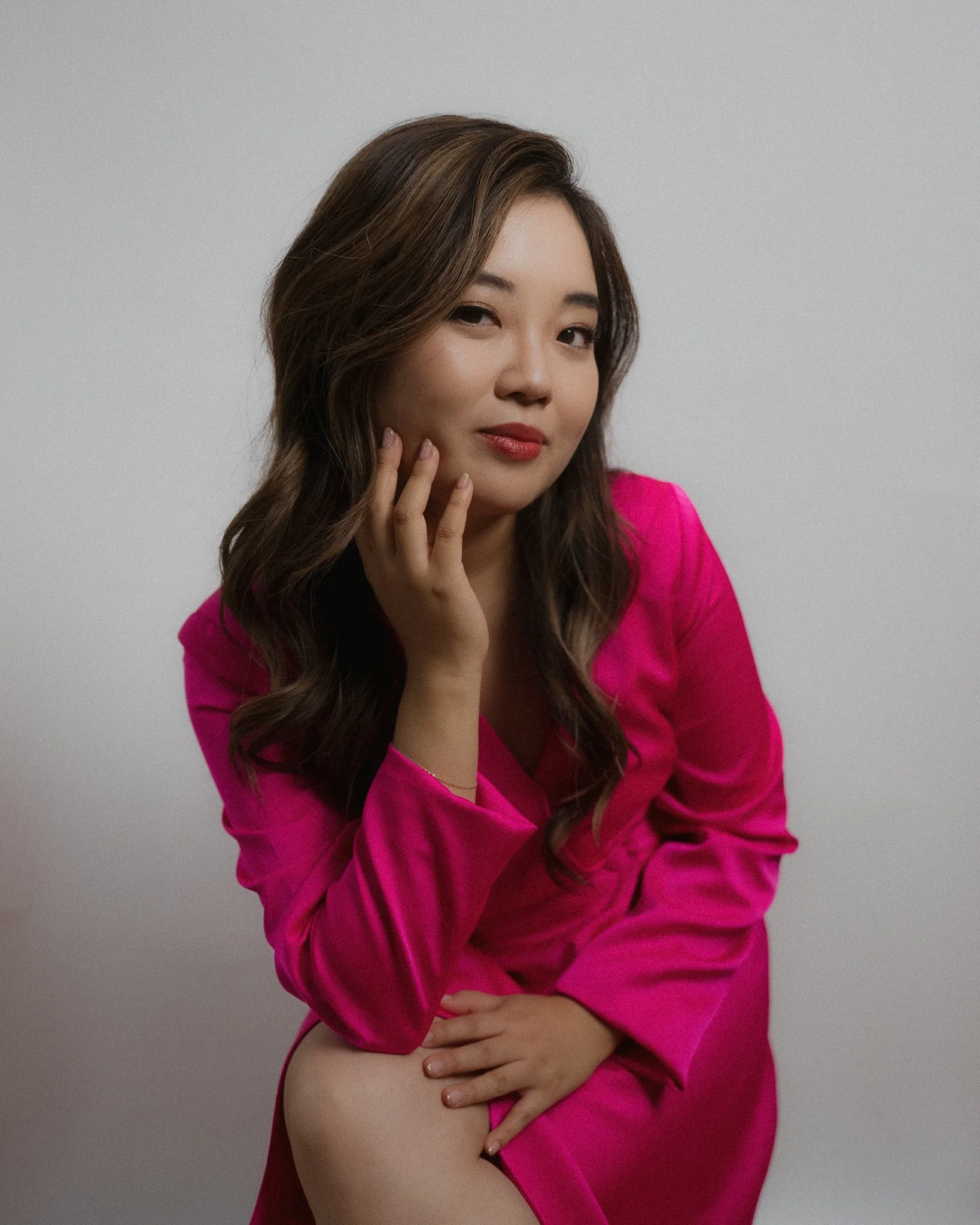 A woman wearing a bright pink satin robe, sitting against a gray background, with one hand resting on her lap and the other touching her face, looking at the camera with a subtle smile.