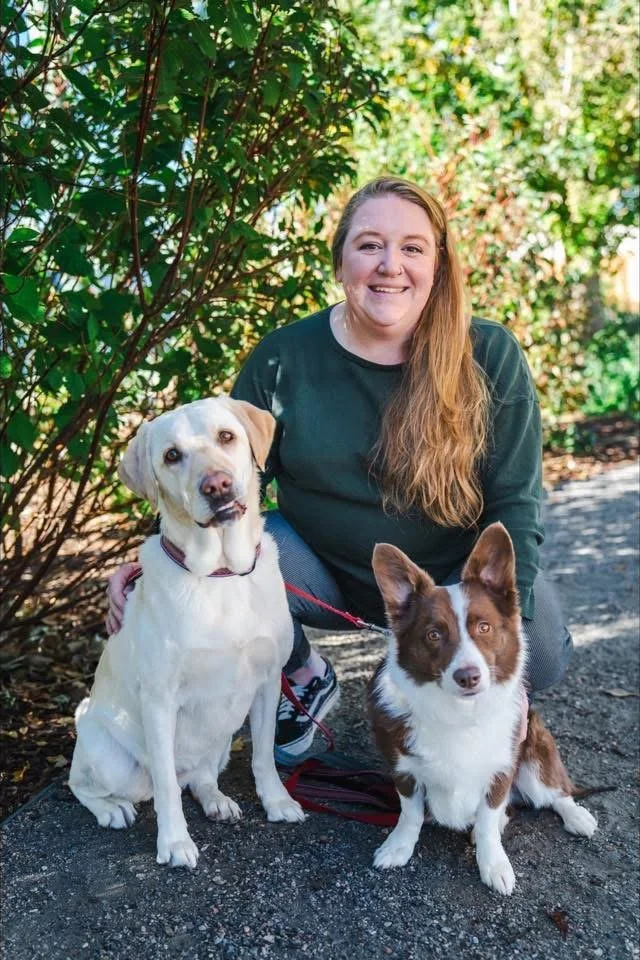 A smiling woman with long red hair kneeling outdoors with two dogs, one white Labrador and one brown and white Border Collie, on a paved trail with green bushes and trees in the background.