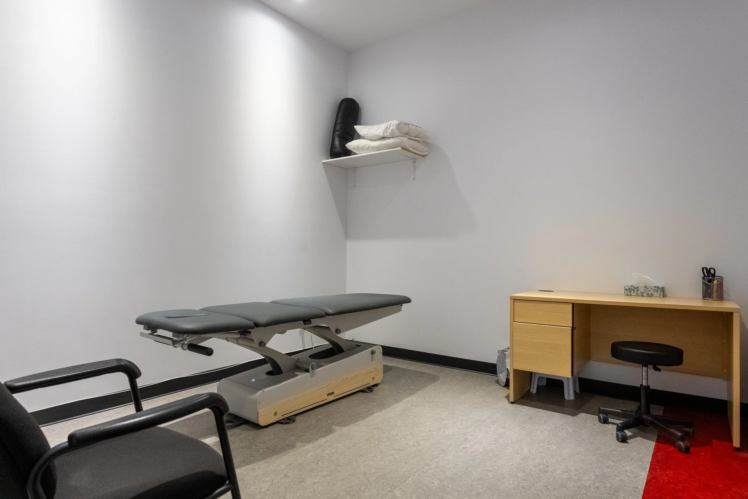 Une pièce d'examen médical avec une table d'examen, un bureau en bois avec des fournitures, un fauteuil noir et une étagère blanche avec des coussins et un rouleau de papier. Sol en moquette grise, mur blanc.