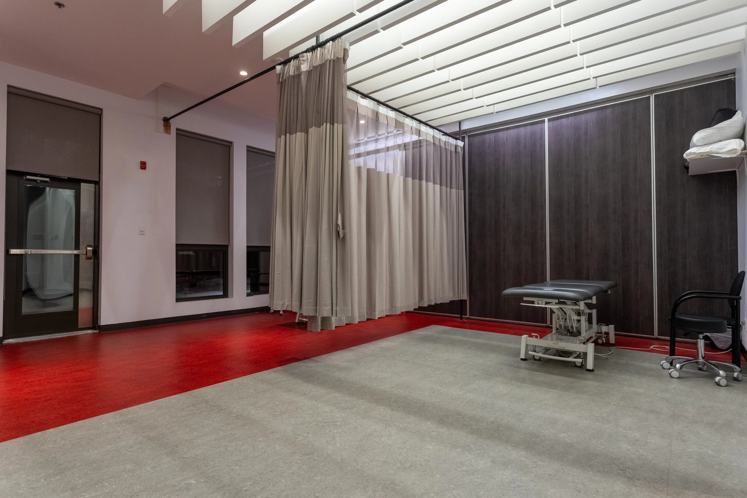 Salle d'examen médical ou clinique avec un lit d'examen, un fauteuil, un rideau de séparation et un mur en bois sombre.