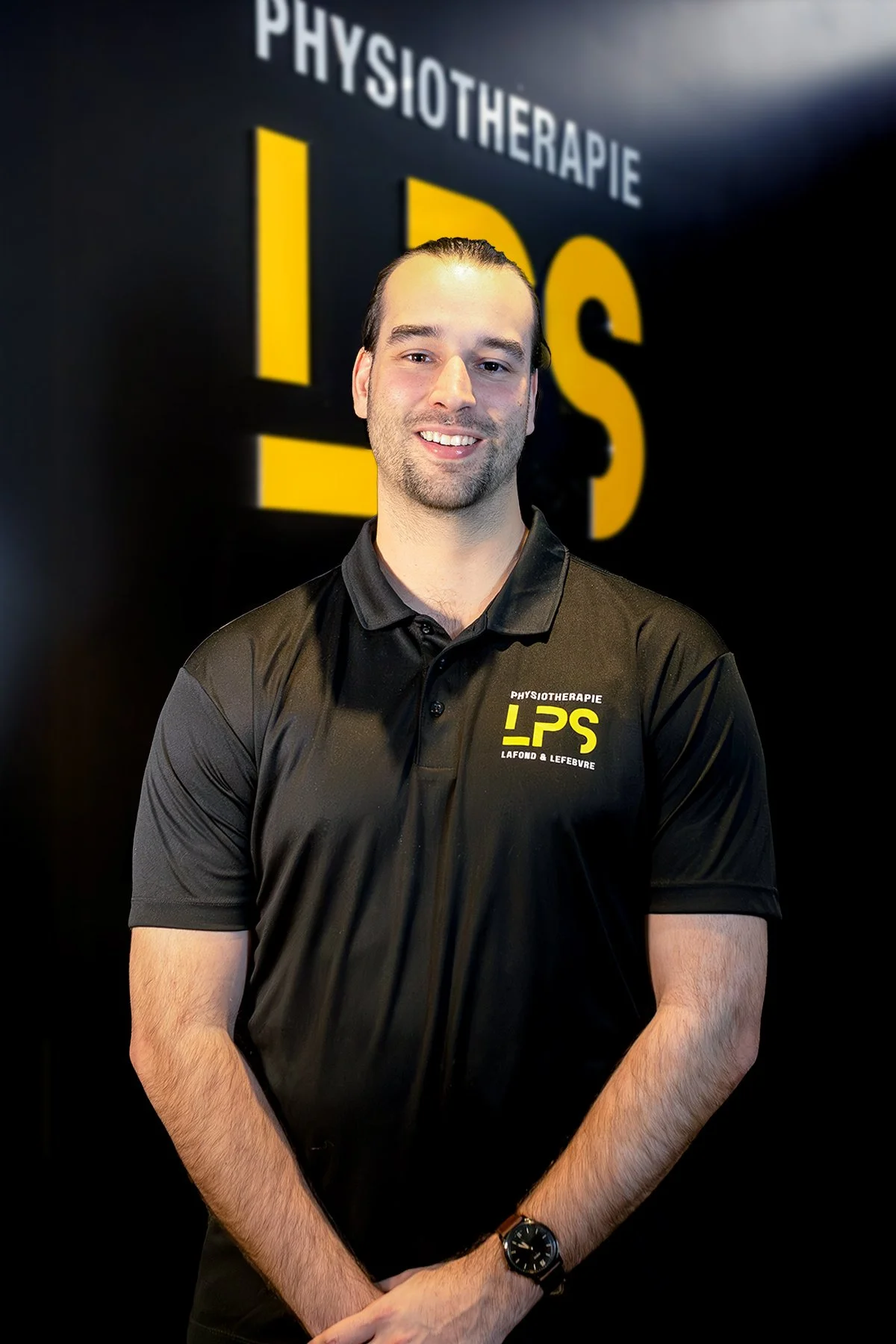 Homme souriant portant un polo noir avec le logo 'Physiothérapie LPS' en jaune, debout devant un mur avec le même logo.