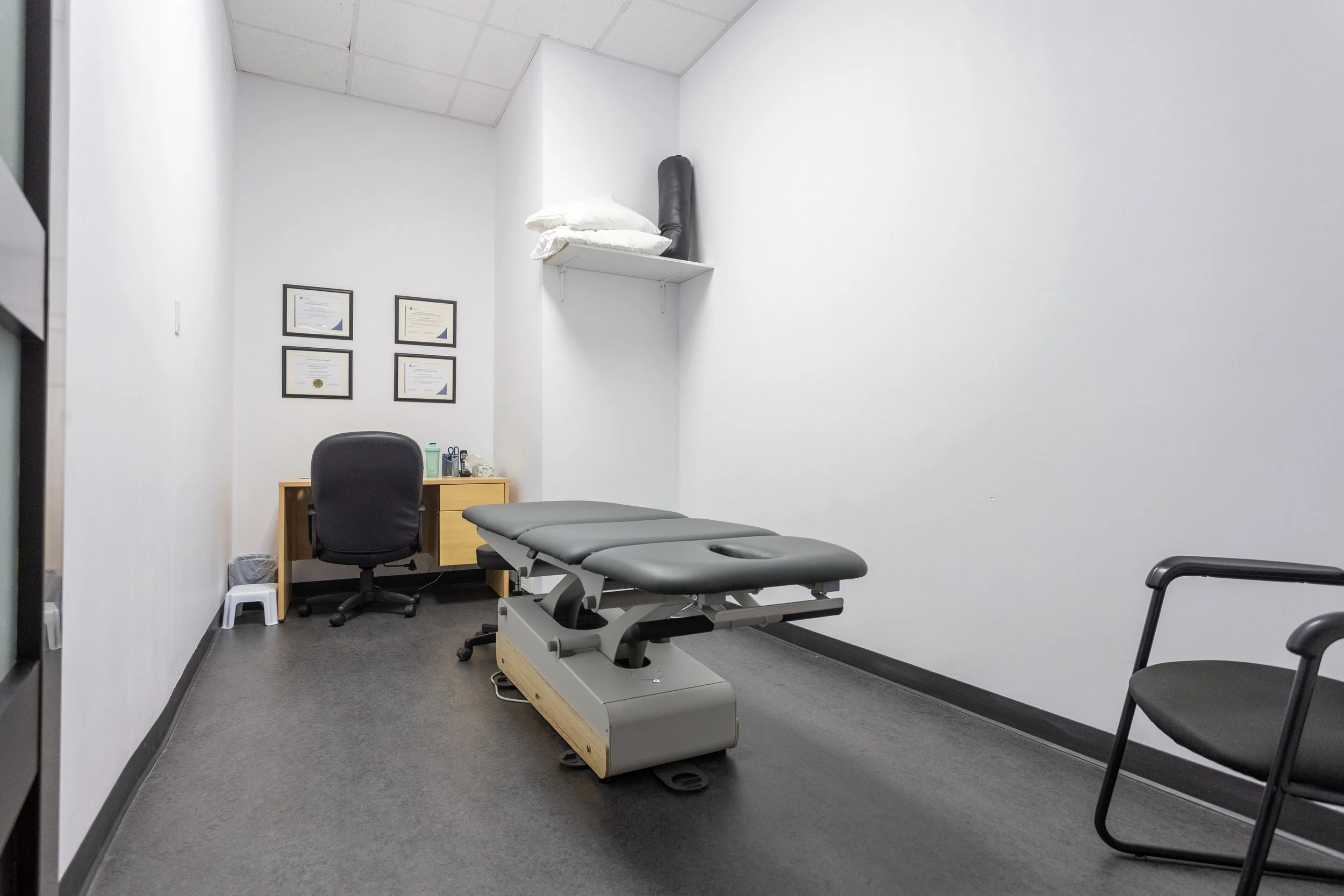 Une salle d'examen médical avec une table de consultation, un bureau avec une chaise, un fauteuil, et des diplômes encadrés au mur.