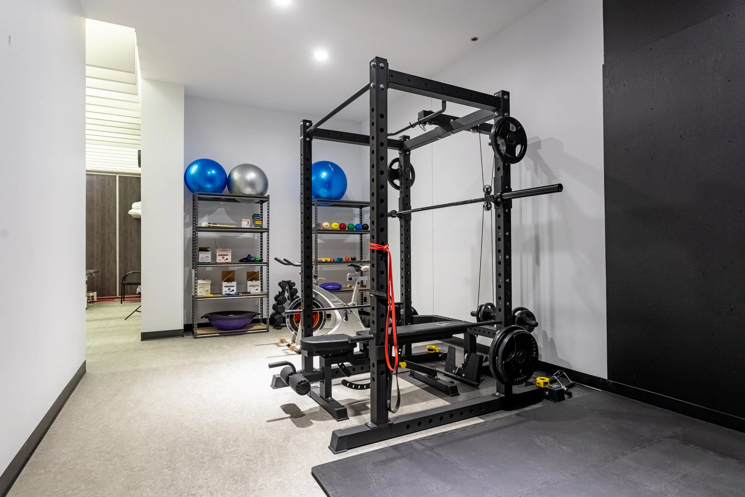 Salle de sport avec un rack d'haltères, une machine à gymnastique, des ballons d'exercice, et des étagères avec des équipements de fitness.