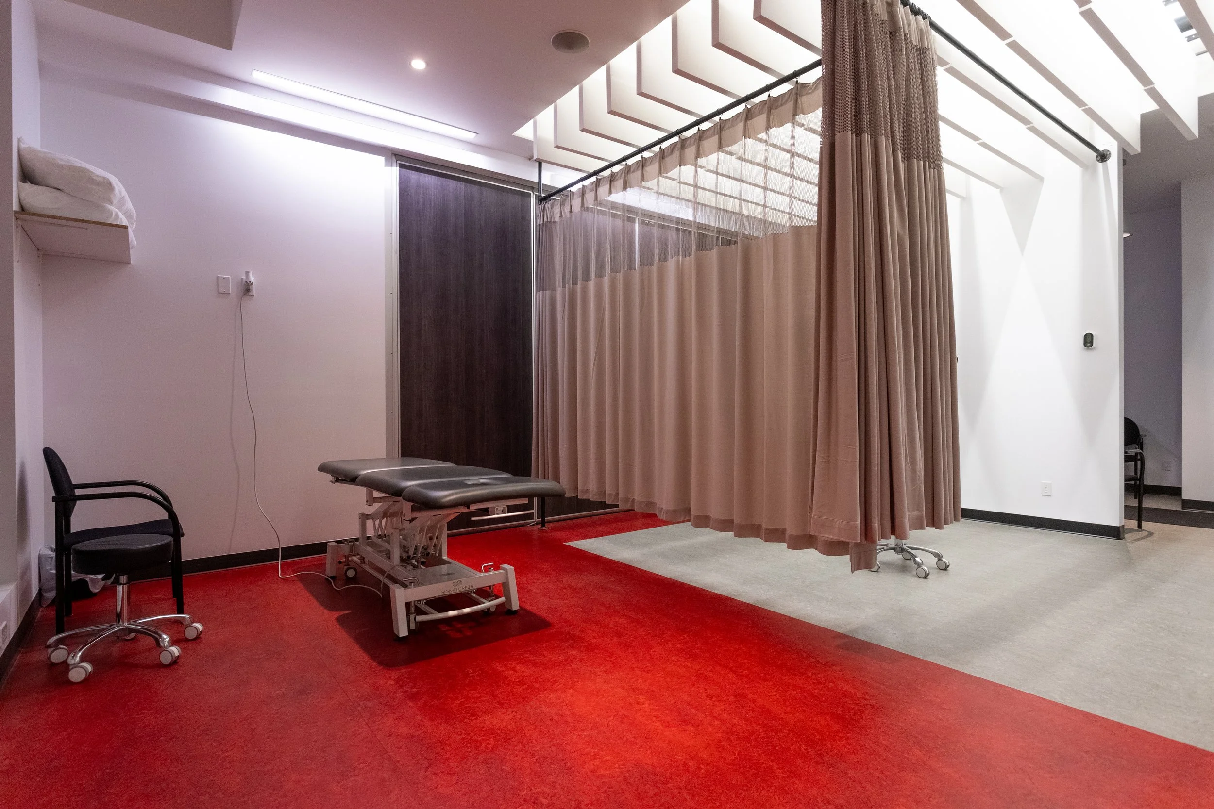 Salle d'examen médical ou thérapeutique avec un rideau pour l'intimité, une table d'examen, deux chaises, et un sol partiellement recouvert de tapis rouge et beige.