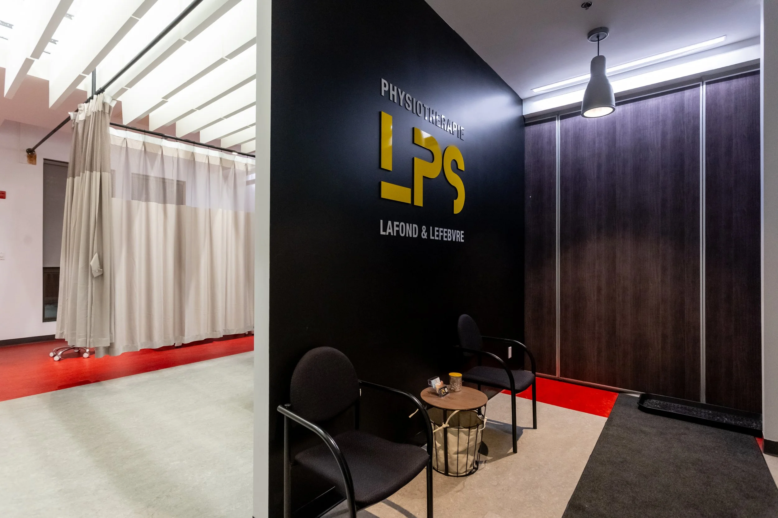 Salle d'attente d'un centre de physiothérapie avec deux chaises, une petite table, et un fauteuil dans un espace moderne avec mûr en bois et un panneau noir avec le nom "Physiotherapie LFSOND & Lefebvre"