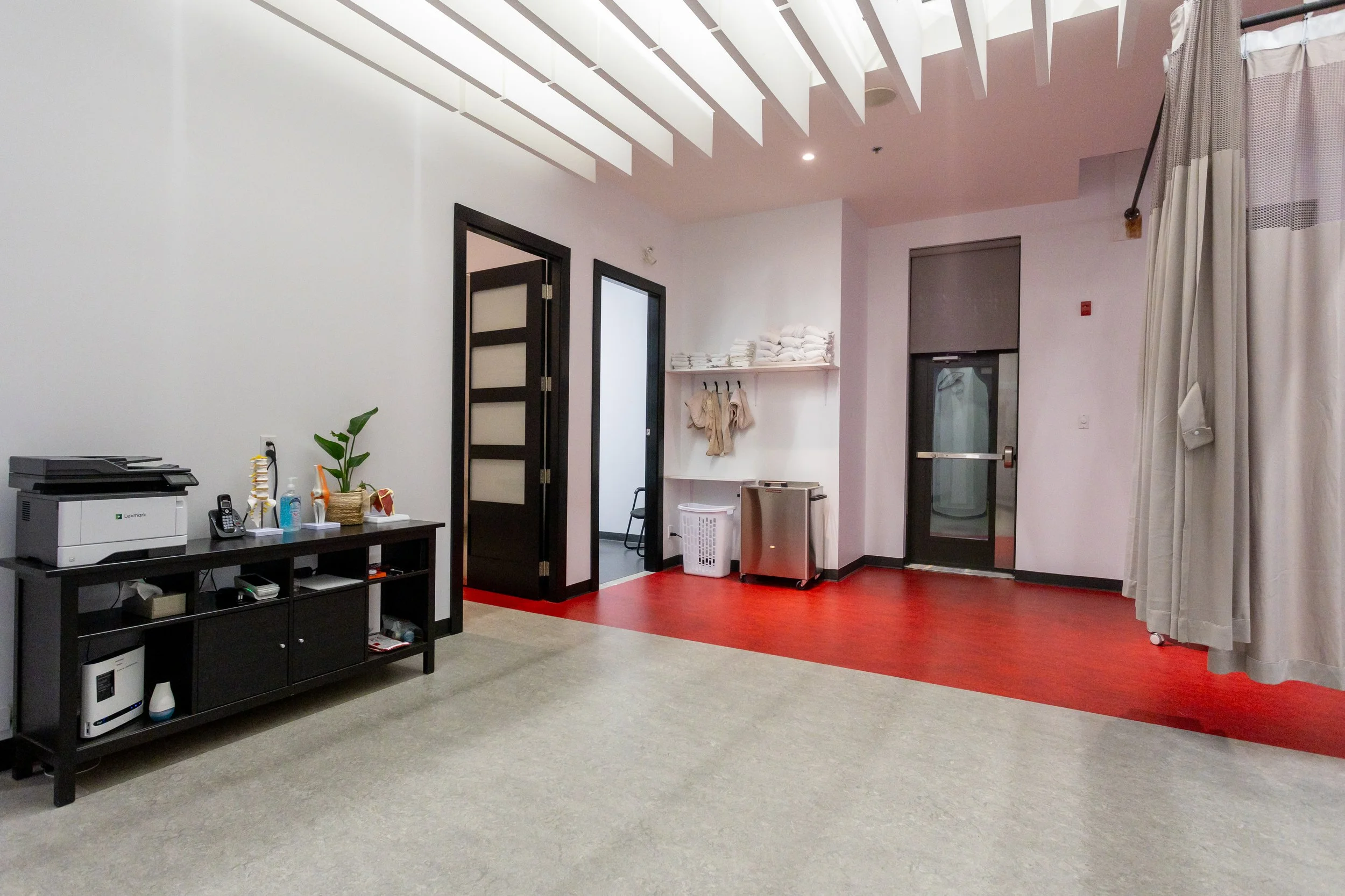 Salle de bain ou spa avec plancher en bois rouge, murs blancs, porte noire, étagère avec serviettes blanches, panier à linge, petits appareils, porte en verre, rideau gris.