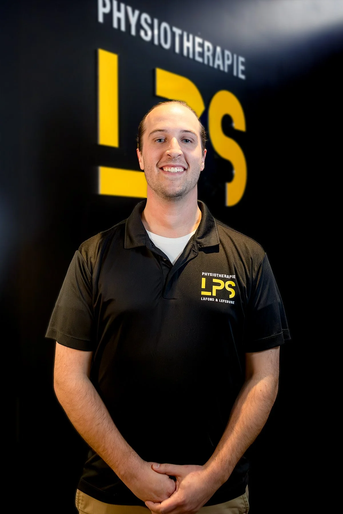 Un homme souriant portant un polo noir avec le logo 'Physiothérapie LaFond & Lefebvre' se tenant devant un fond noir avec le même logo en grand.