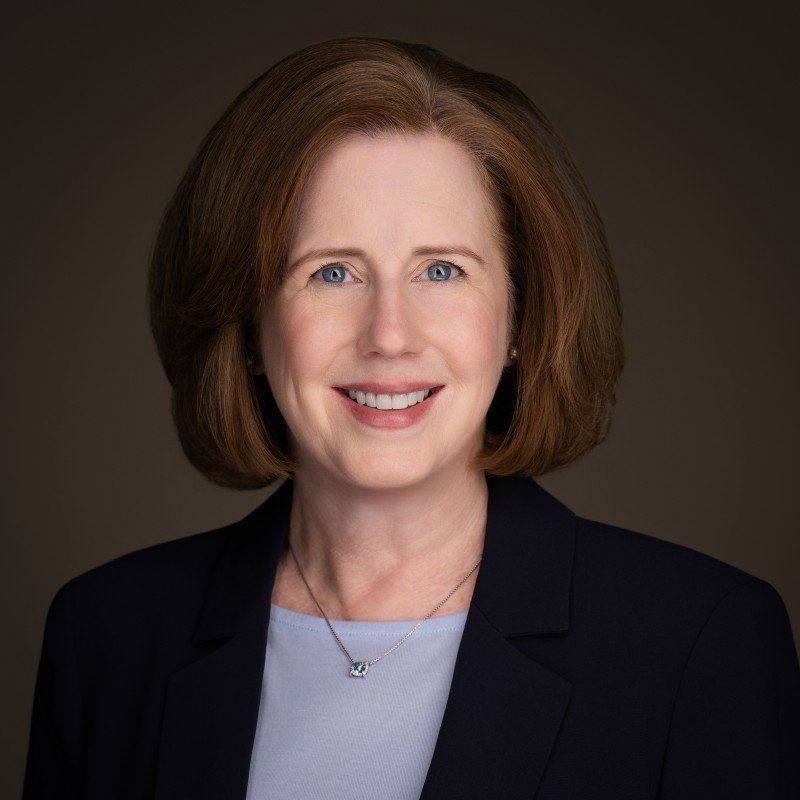 Close-up photograph of a smiling woman with shoulder-length brown hair, wearing pearl earrings and a dark red top, outdoors with green trees and sunlight in the background.