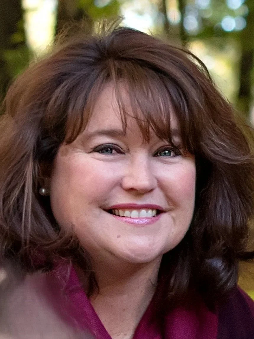 Close-up photograph of a smiling woman with shoulder-length brown hair, wearing pearl earrings and a dark red top, outdoors with green trees and sunlight in the background.