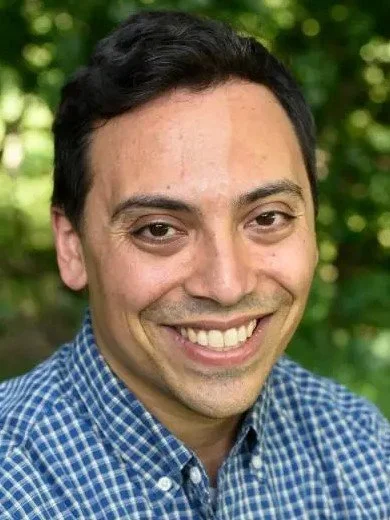 A smiling bald man wearing glasses and a navy blazer, in front of a bookshelf filled with books.