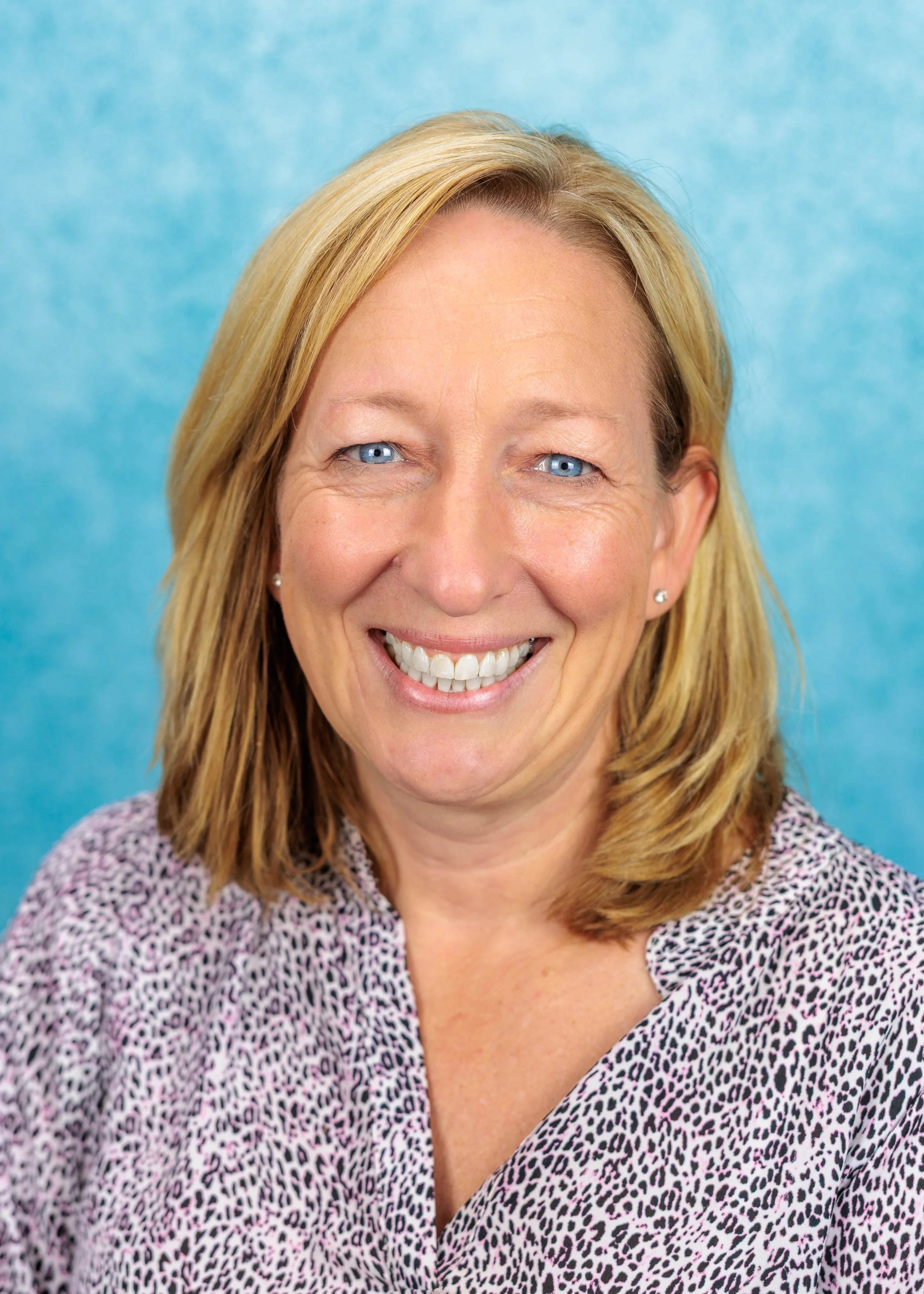 Smiling woman with short blond hair, wearing a yellow top, standing in front of a patterned tapestry with birds and floral designs.