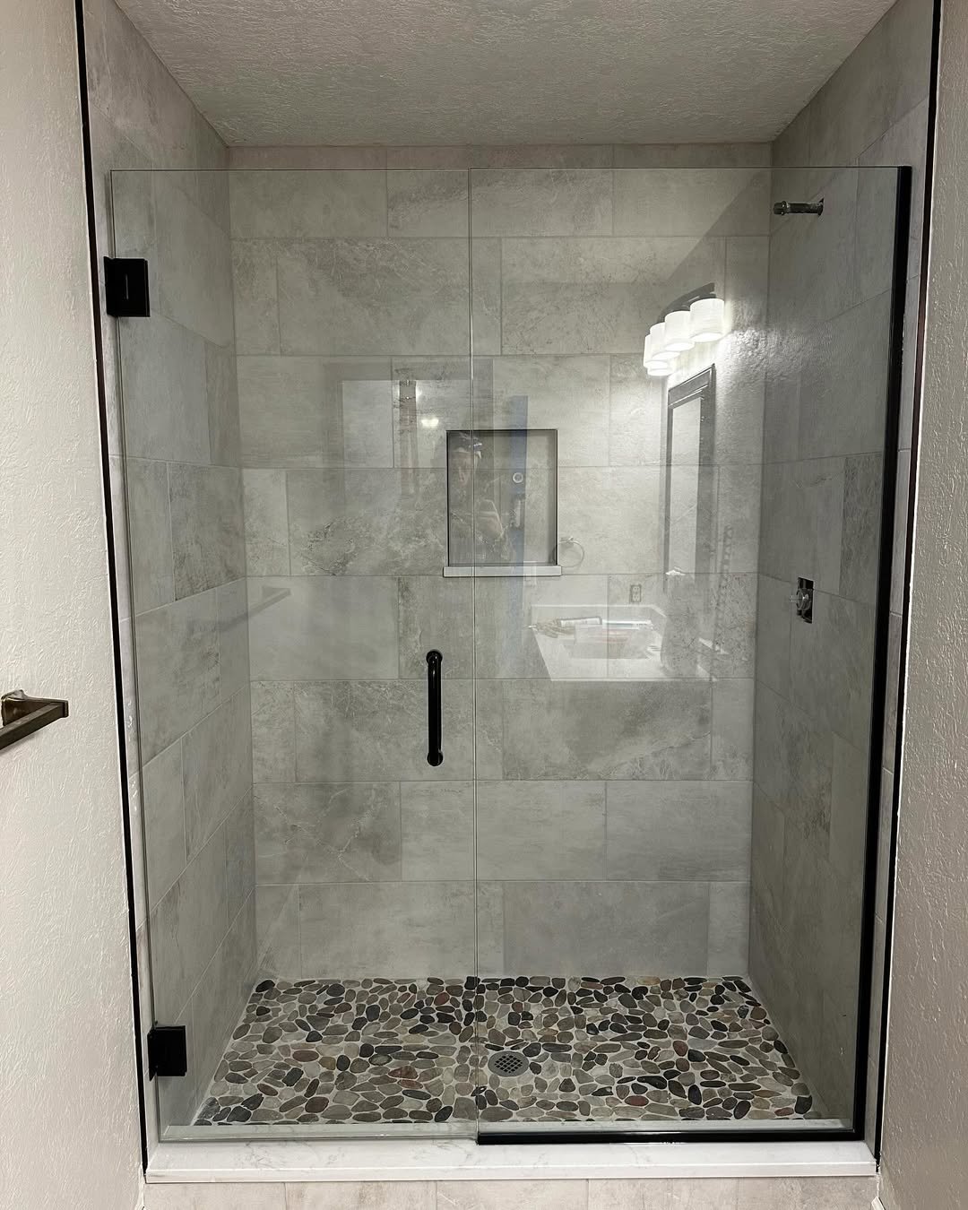 A glass-enclosed shower with tiled walls and pebble stone flooring, a built-in niche, and a door handle.
