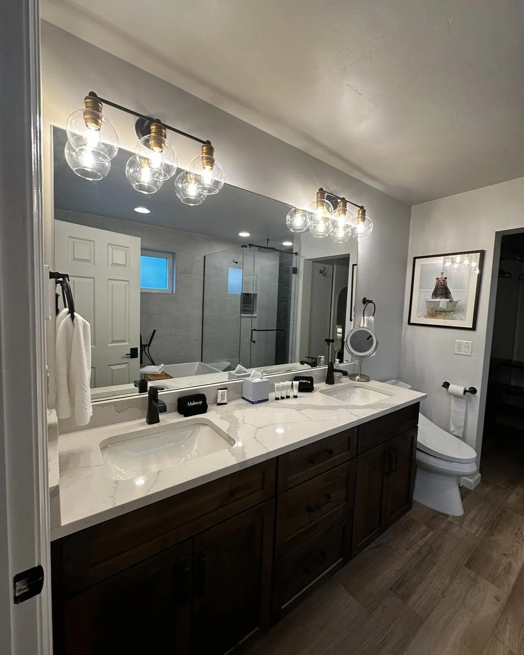Modern bathroom with double sink vanity, large mirror, wooden cabinets, marble countertop, wall-mounted light fixtures, frameless glass shower, framed art, and toilet.