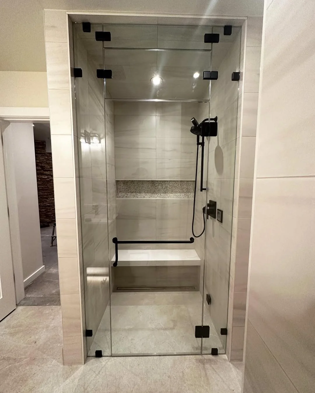 A modern walk-in shower with clear glass doors, black fixtures, a built-in bench, and a rain shower head, surrounded by light-colored tiles.