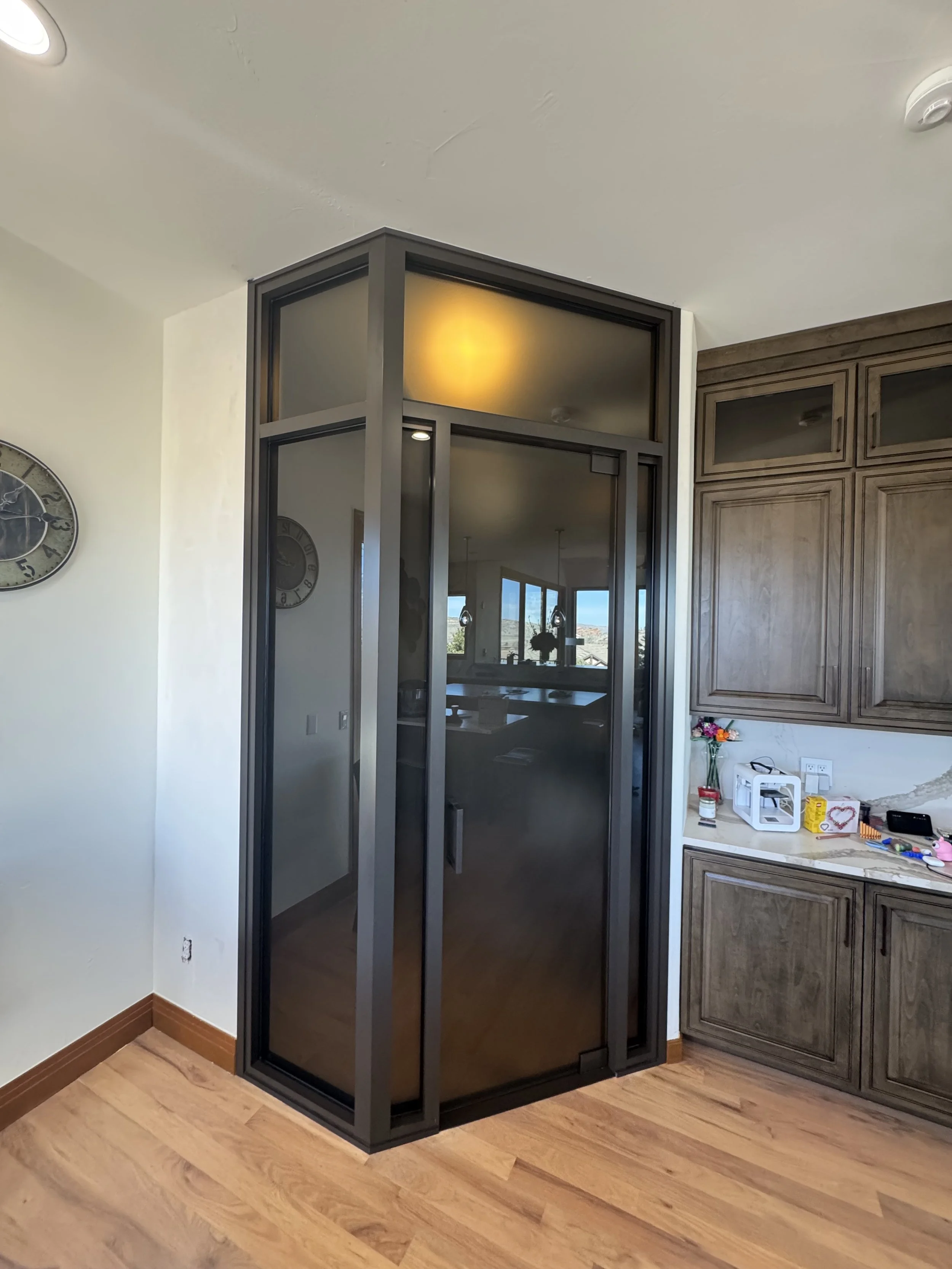 Black framed glass corner shower enclosure in a kitchen area with wooden cabinets and hardwood floors.