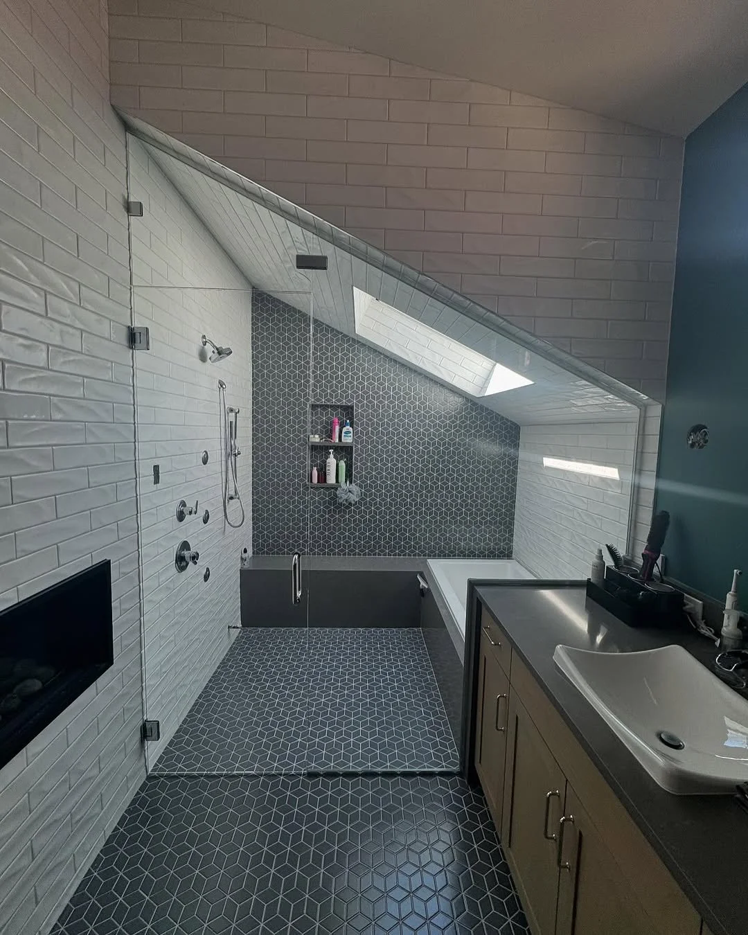 Modern bathroom with sloped ceiling and skylight, featuring a walk-in shower with glass door, built-in niche, black and white hexagonal and rectangular tiles, a bathtub, and a vanity with sink and toiletries.