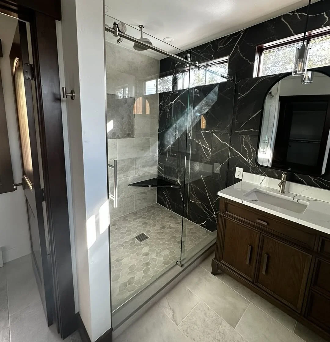 Modern bathroom with a glass shower enclosure, black marble walls, hexagonal tile floor in the shower, and a wooden vanity with a white countertop and a rectangular sink.