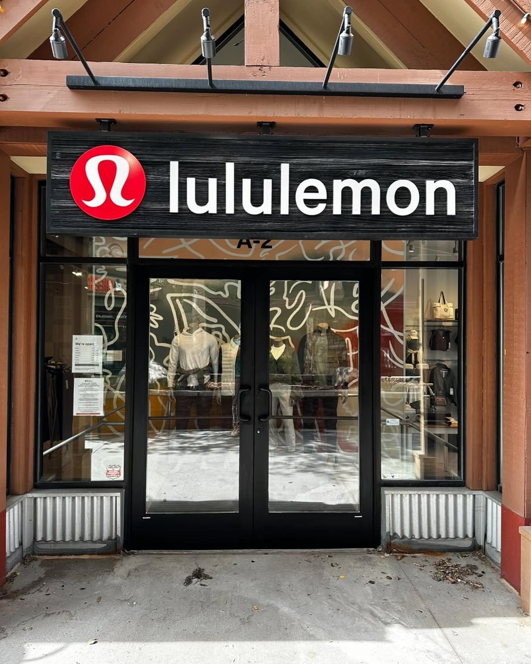 Exterior view of a Lululemon store with black signboard and white logo, glass doors, and window display of mannequins wearing athletic clothing.