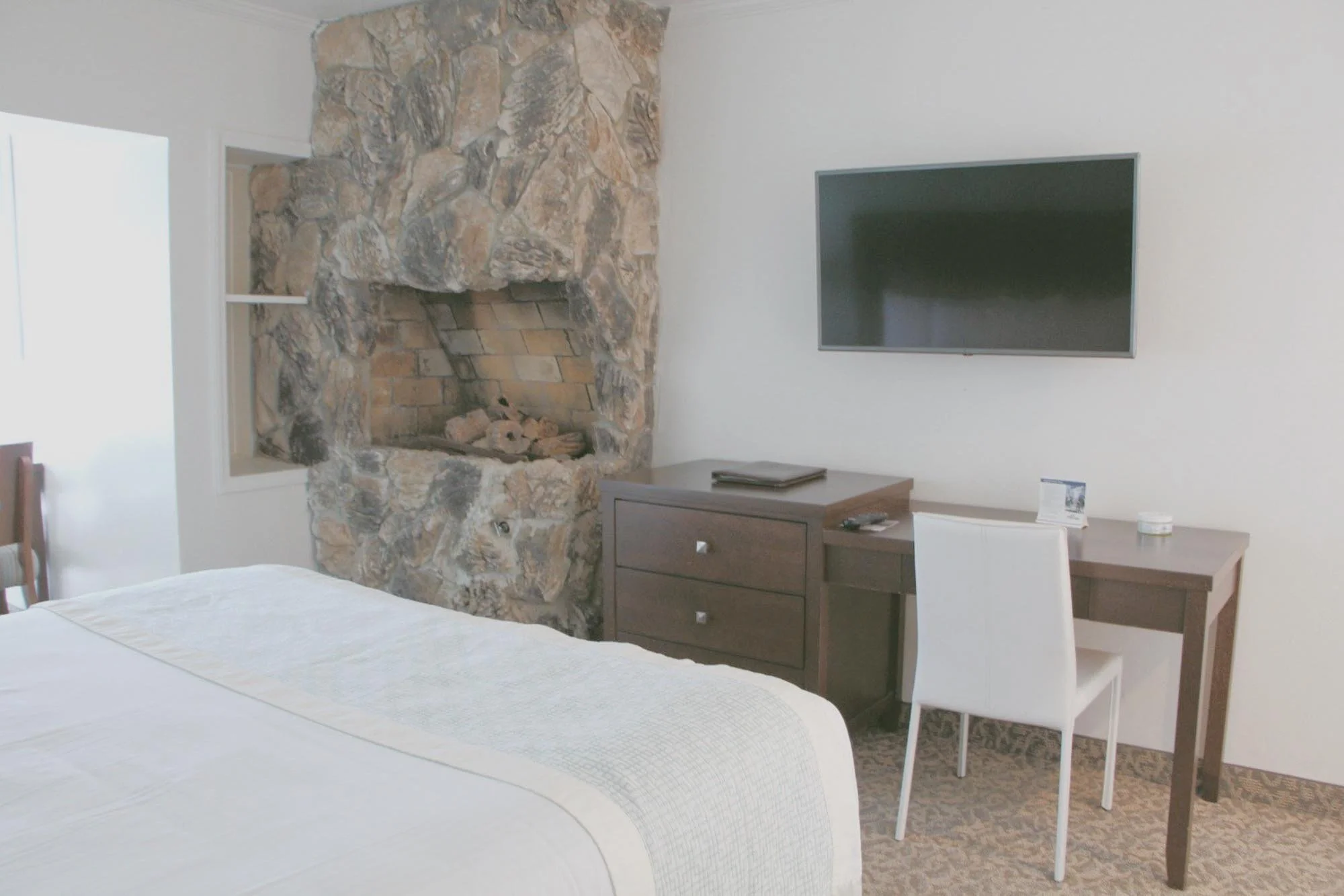 Hotel room with a stone fireplace, a wall-mounted flat-screen TV, a wooden dresser, a white chair, and part of a bed with white linens.
