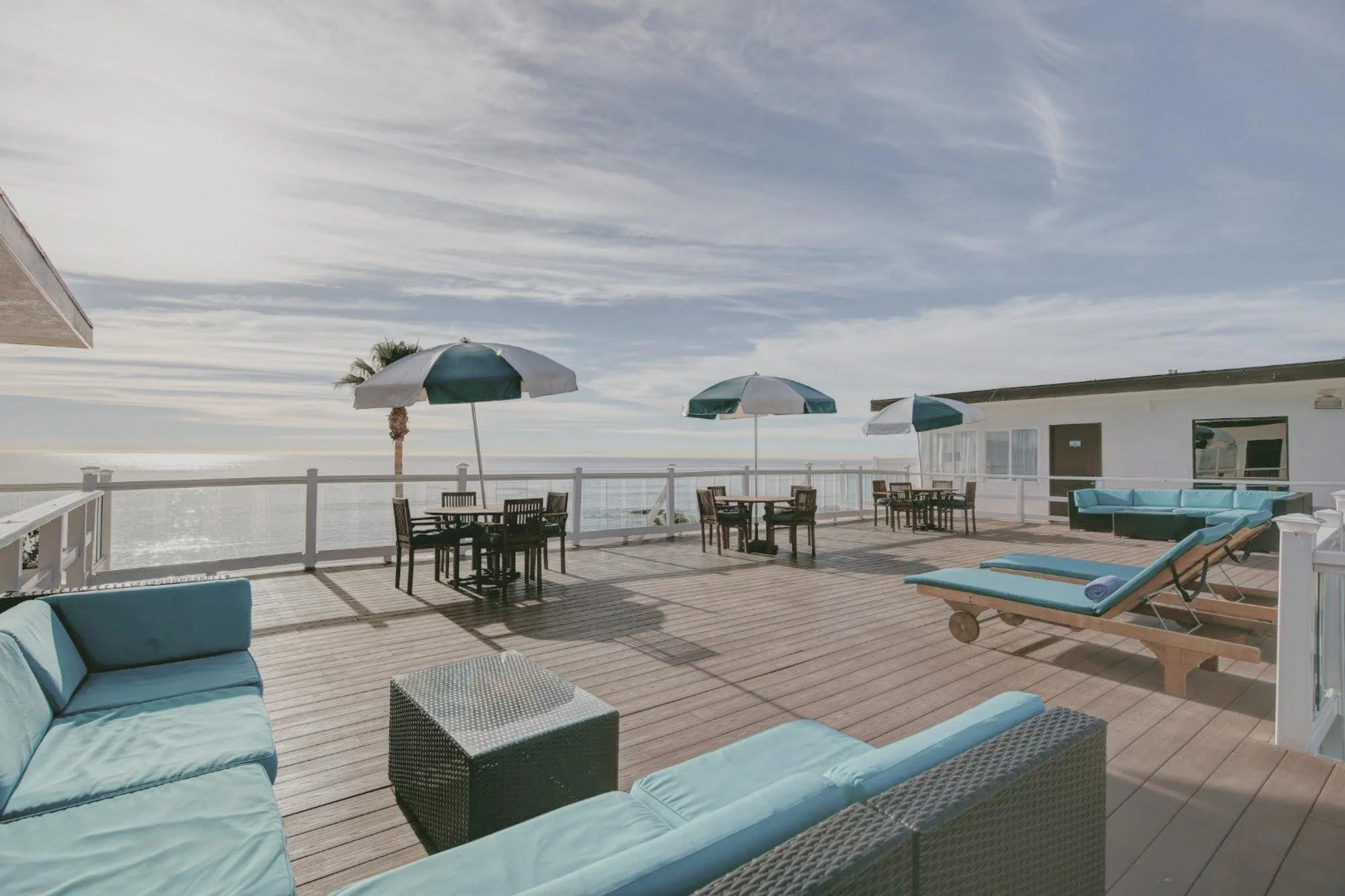 Beachfront outdoor patio with blue cushioned lounge chairs, wooden deck, white railing, and green and white umbrellas, overlooking the ocean under a partly cloudy sky.