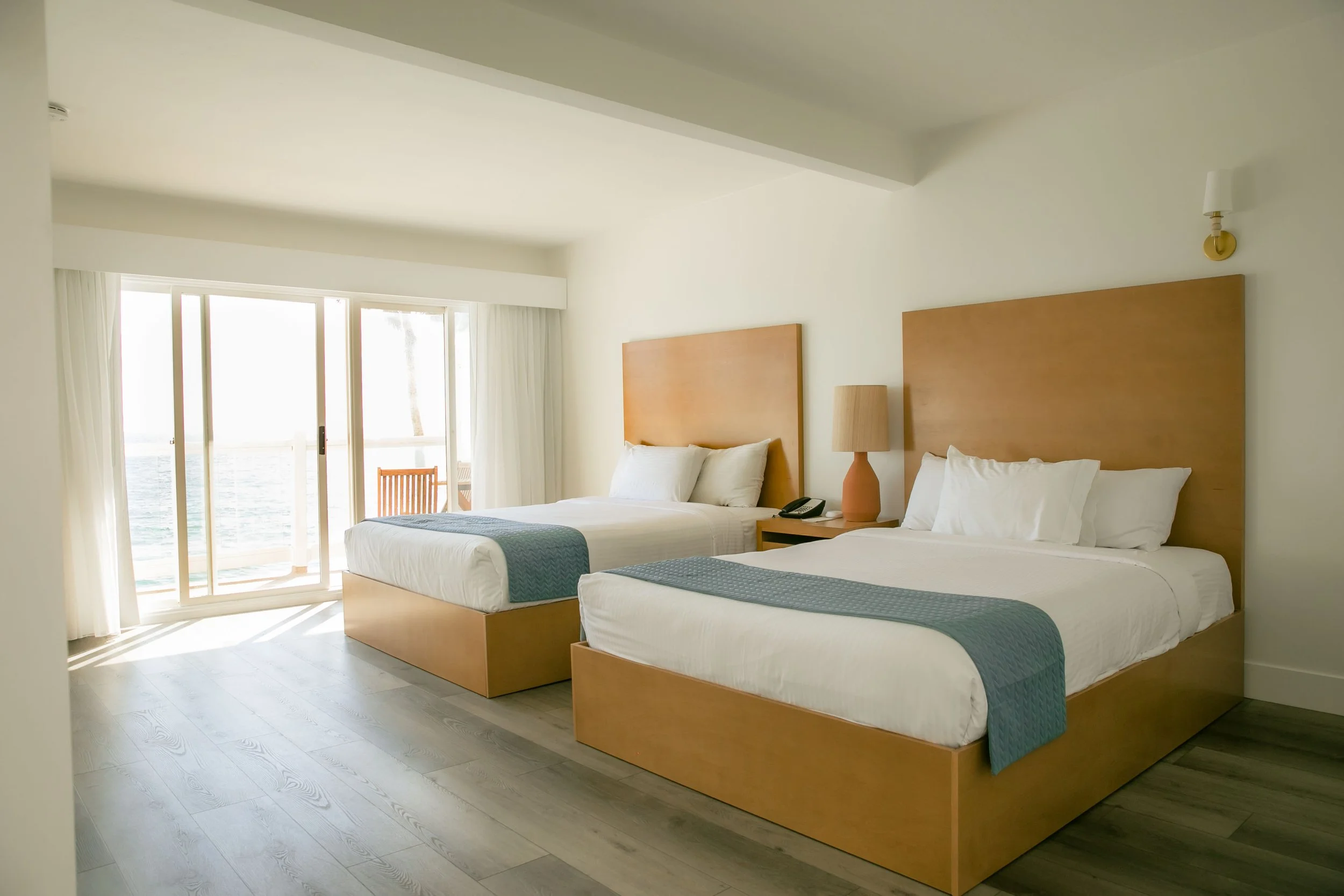 Hotel room with two double beds, a bedside table with a lamp, a telephone, and a curtain-covered sliding door leading to a balcony with a view of the ocean.