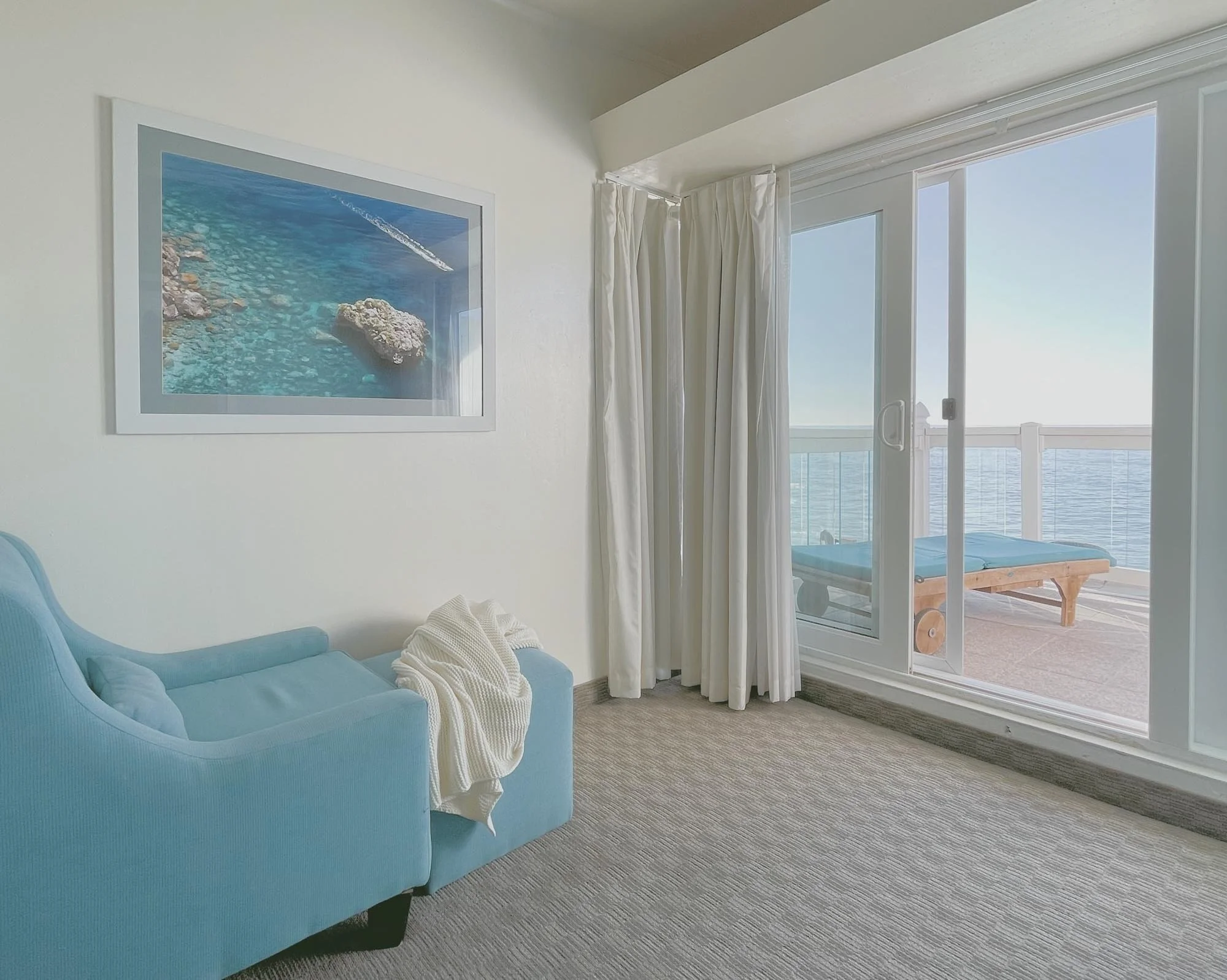 Hotel room with blue armchair, white blanket, wall art of ocean scene, glass sliding door leading to balcony with ocean view and lounge chair.