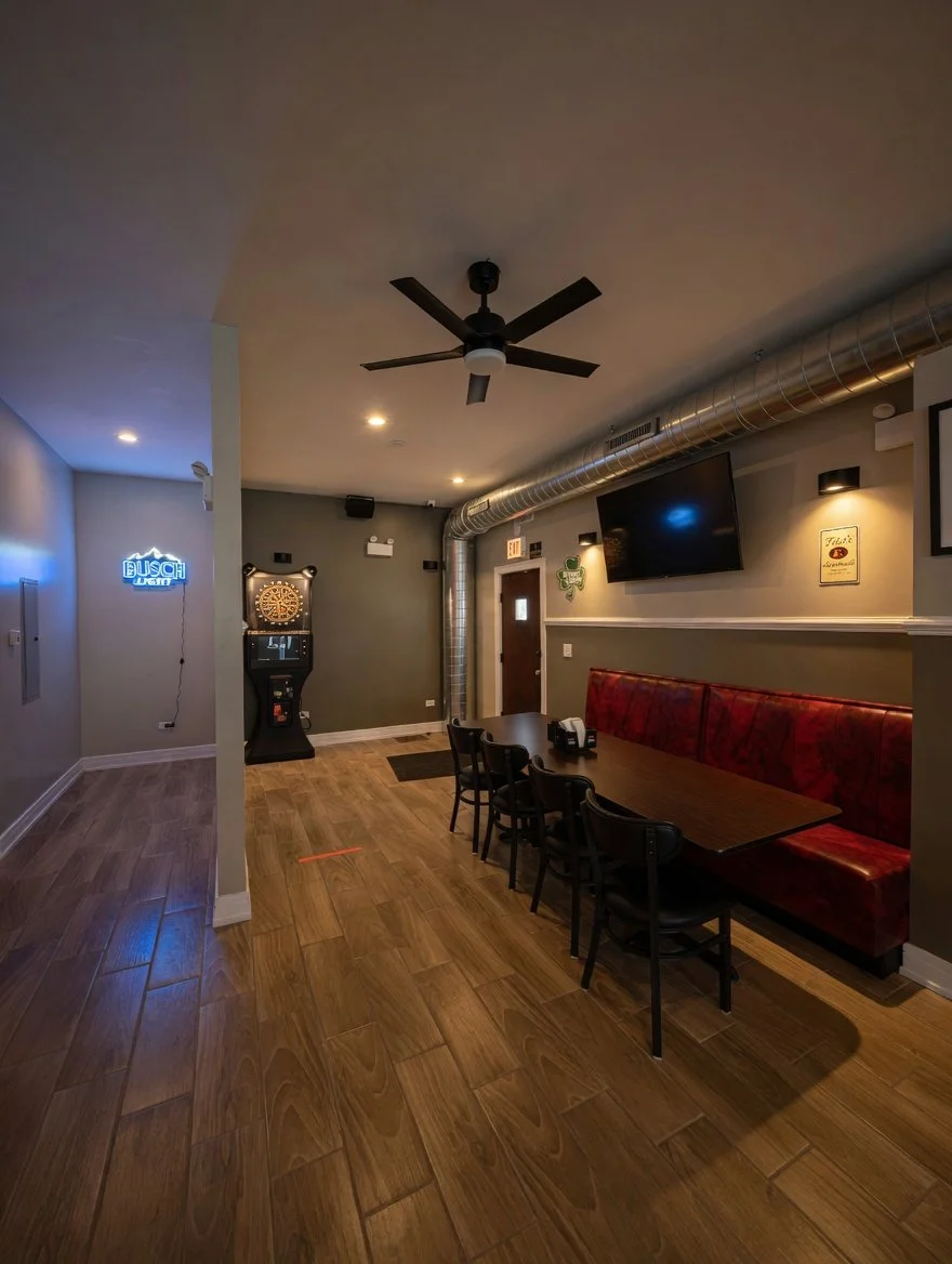 Interior of a bar or restaurant with a long wooden table, black chairs, a red upholstered bench, a dartboard, a TV on the wall, a ceiling fan, and dim lighting.