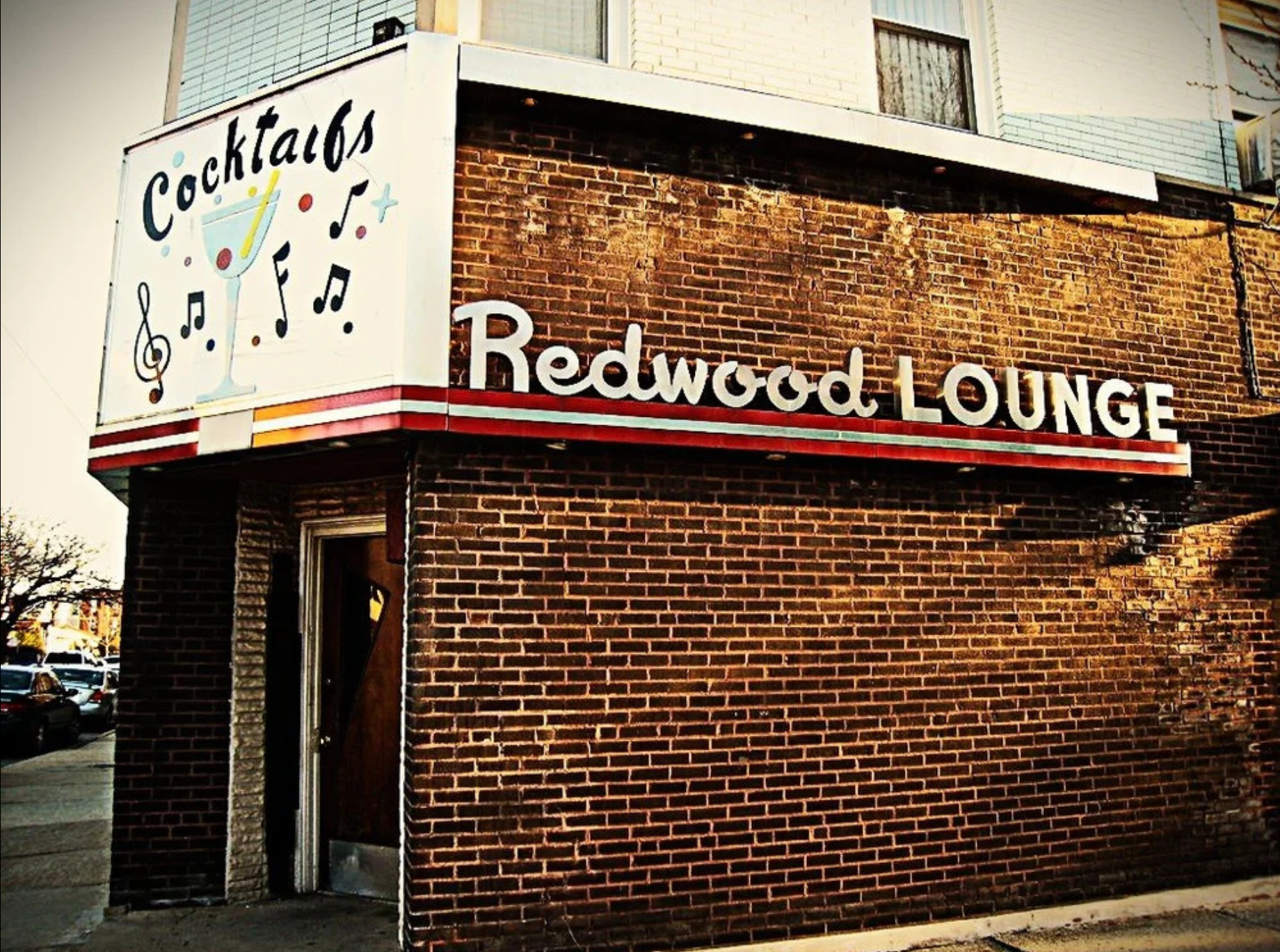 Corner of a brick building with a white sign that reads 'Redwood Lounge' and a second sign that features a cocktail glass, music notes, and the word 'cocktails'.