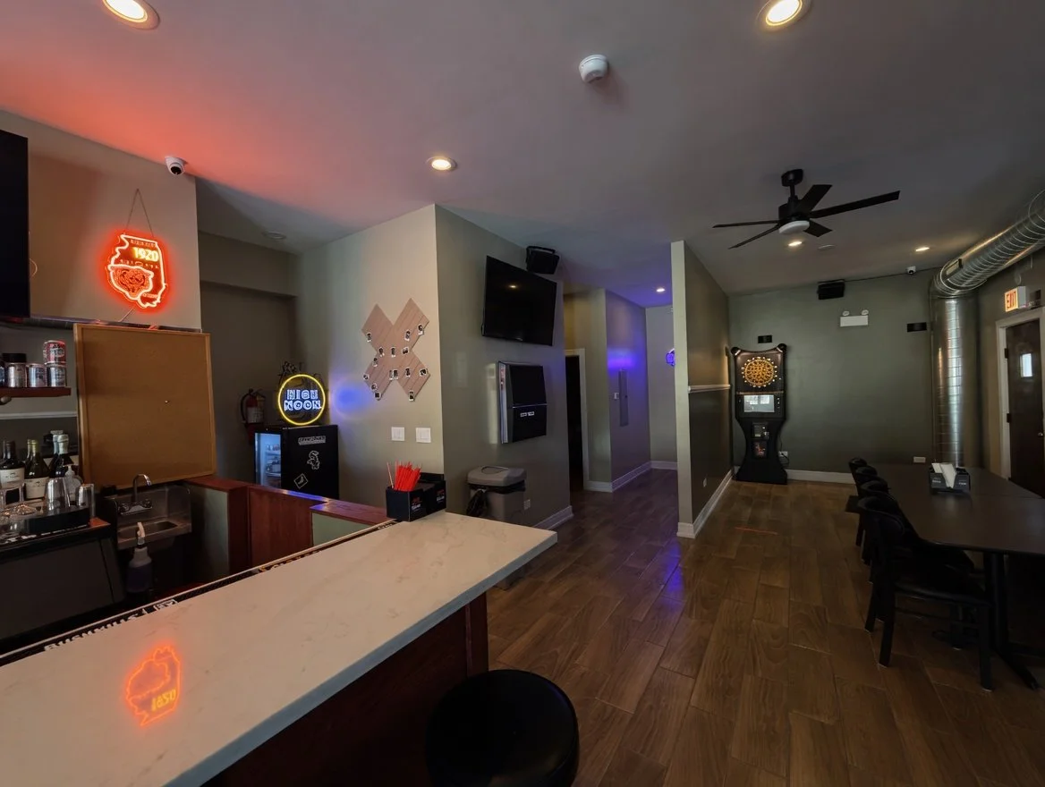Interior view of a bar or game room with a bar counter, neon signs, a dartboard, multiple TVs, and seating area, featuring wood flooring and ceiling fans.