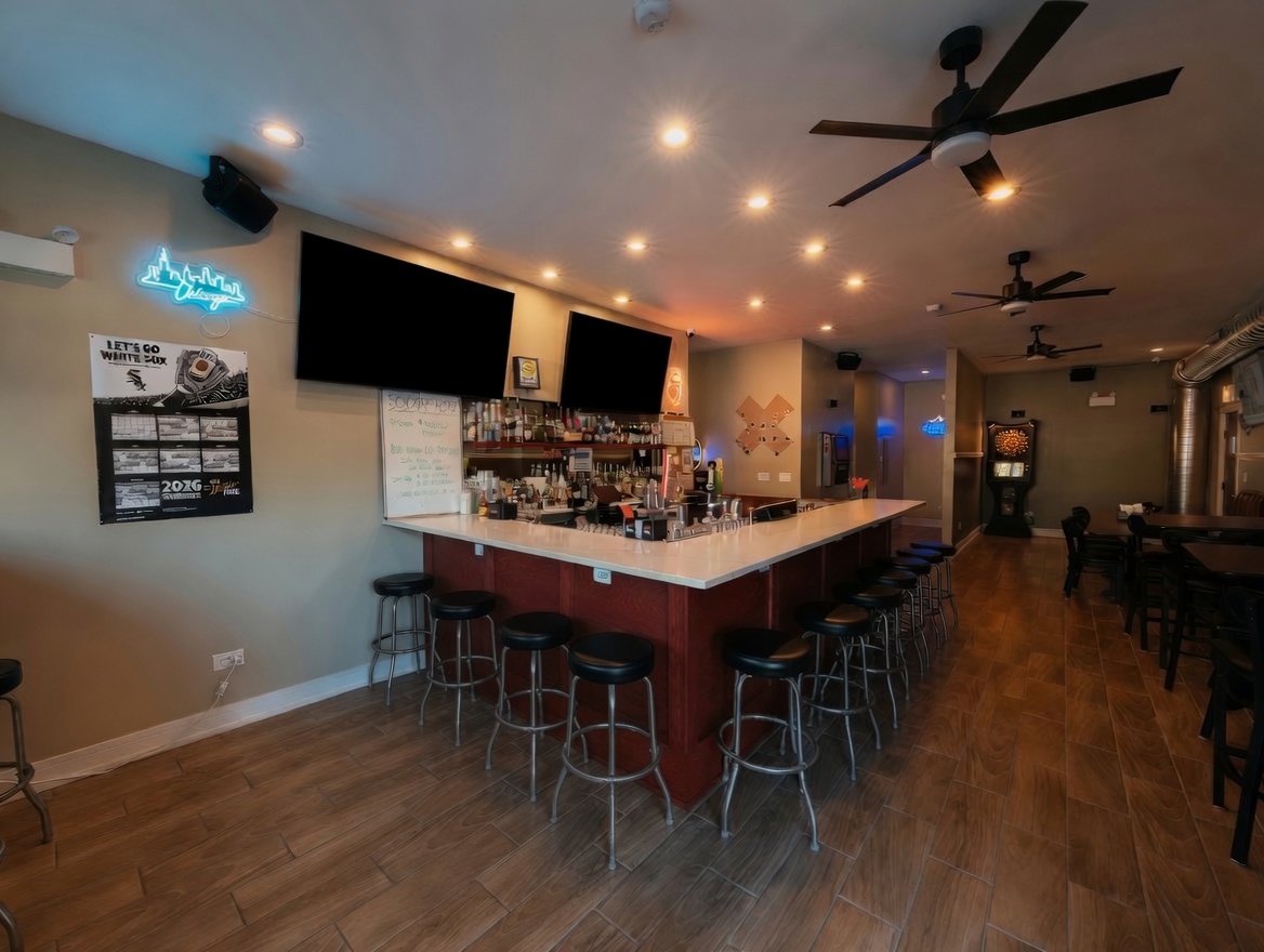 Interior of a bar with a white counter, barstools, two large TVs on the wall, ceiling fans, and a dartboard in the background.