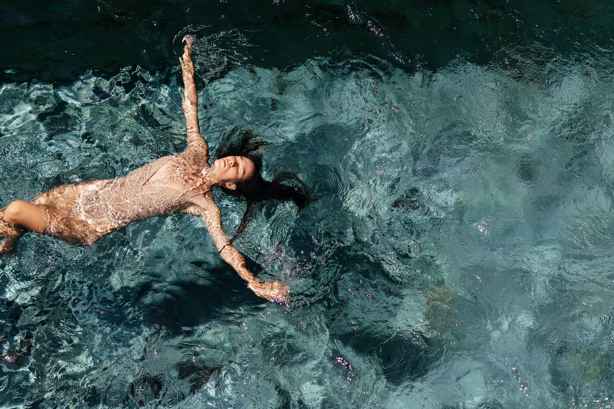 Woman wearing a tan bathing suit is floating in a pool with her eyes closed looking relaxed.