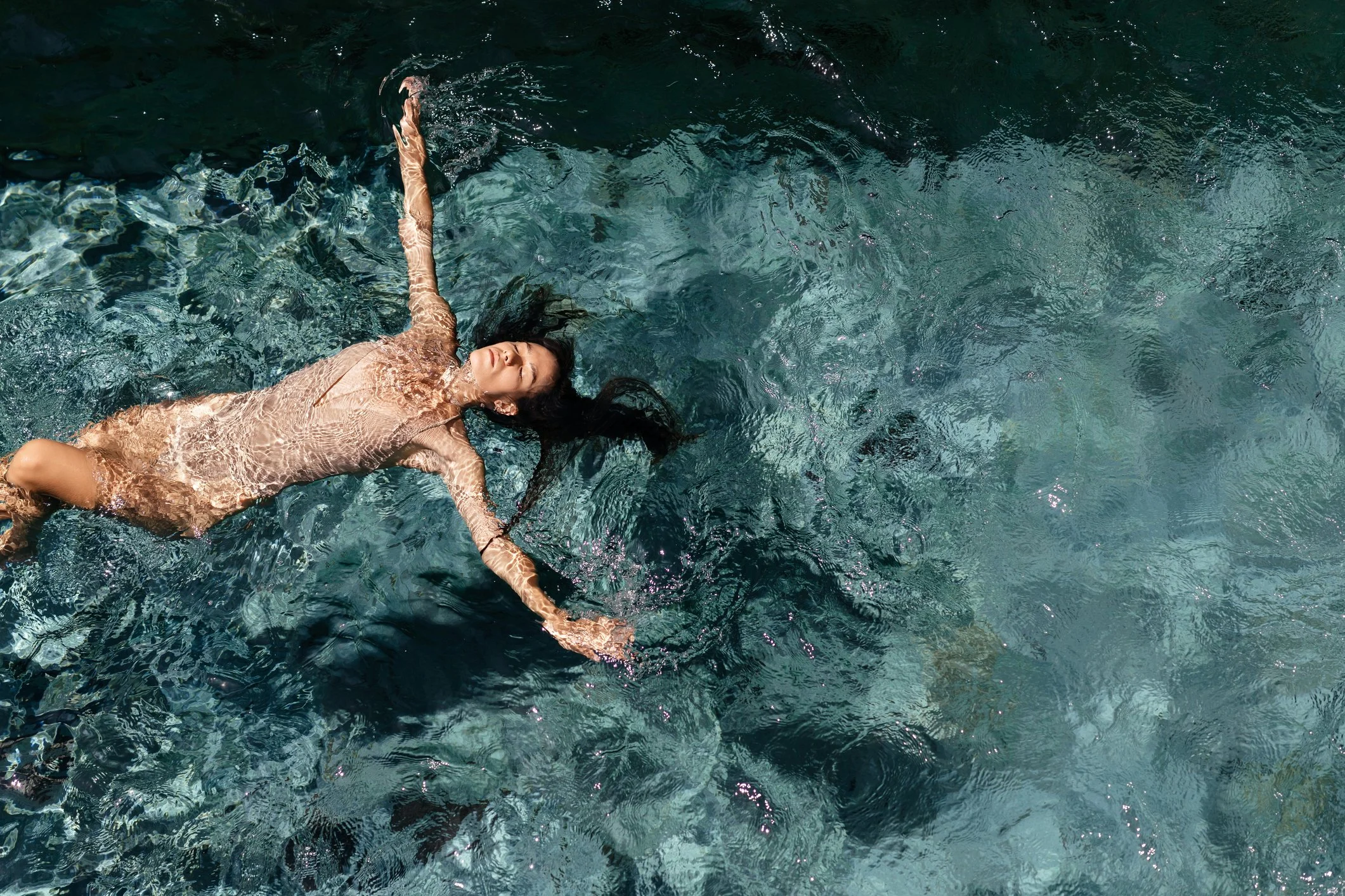 A woman with long dark hair floating on her back in a clear, greenish-blue body of water, with her arms outstretched and eyes closed.