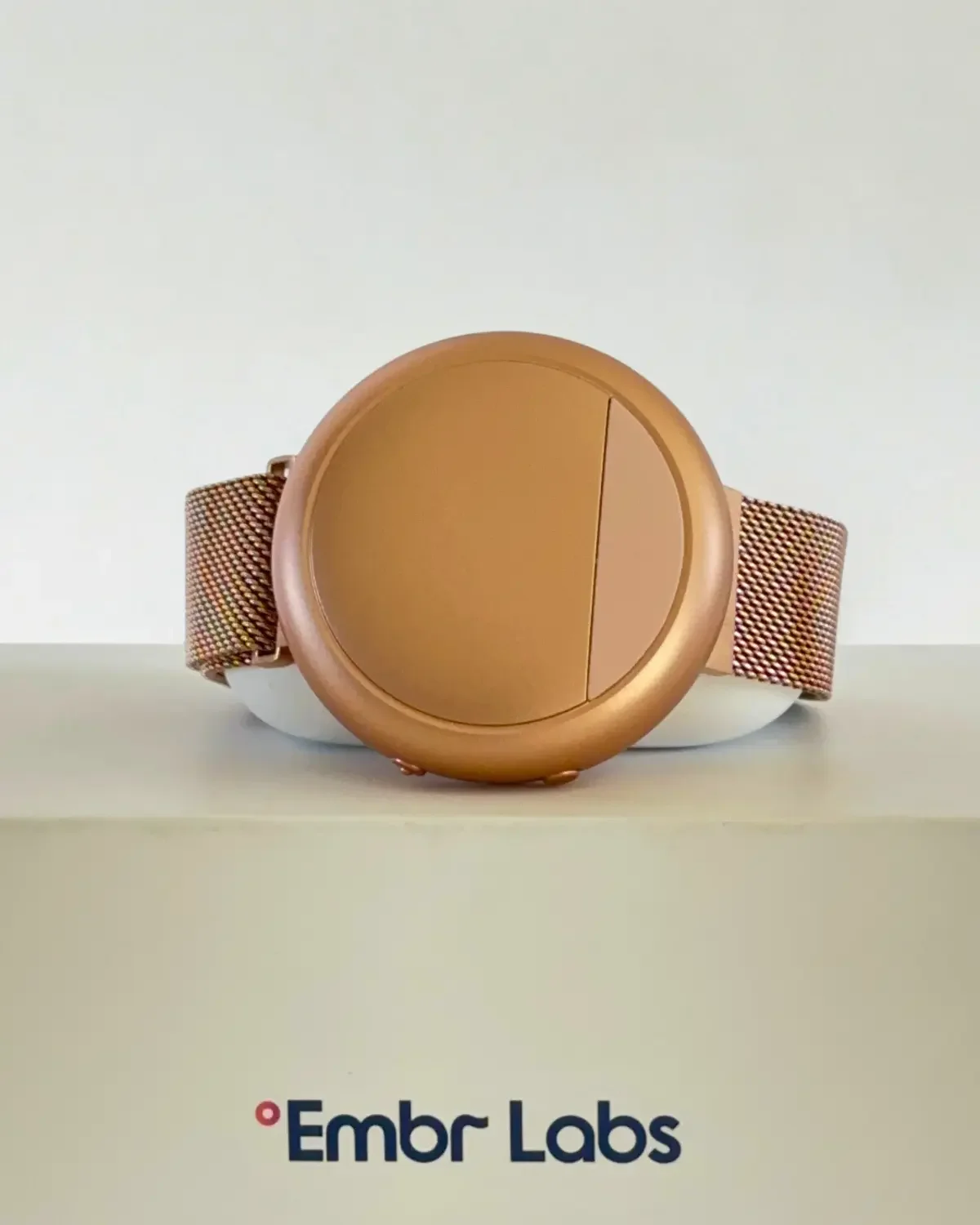 A rose gold smartwatch with a band made of mesh metal, placed on a white box with label 'Embr Labs' against a light white background.