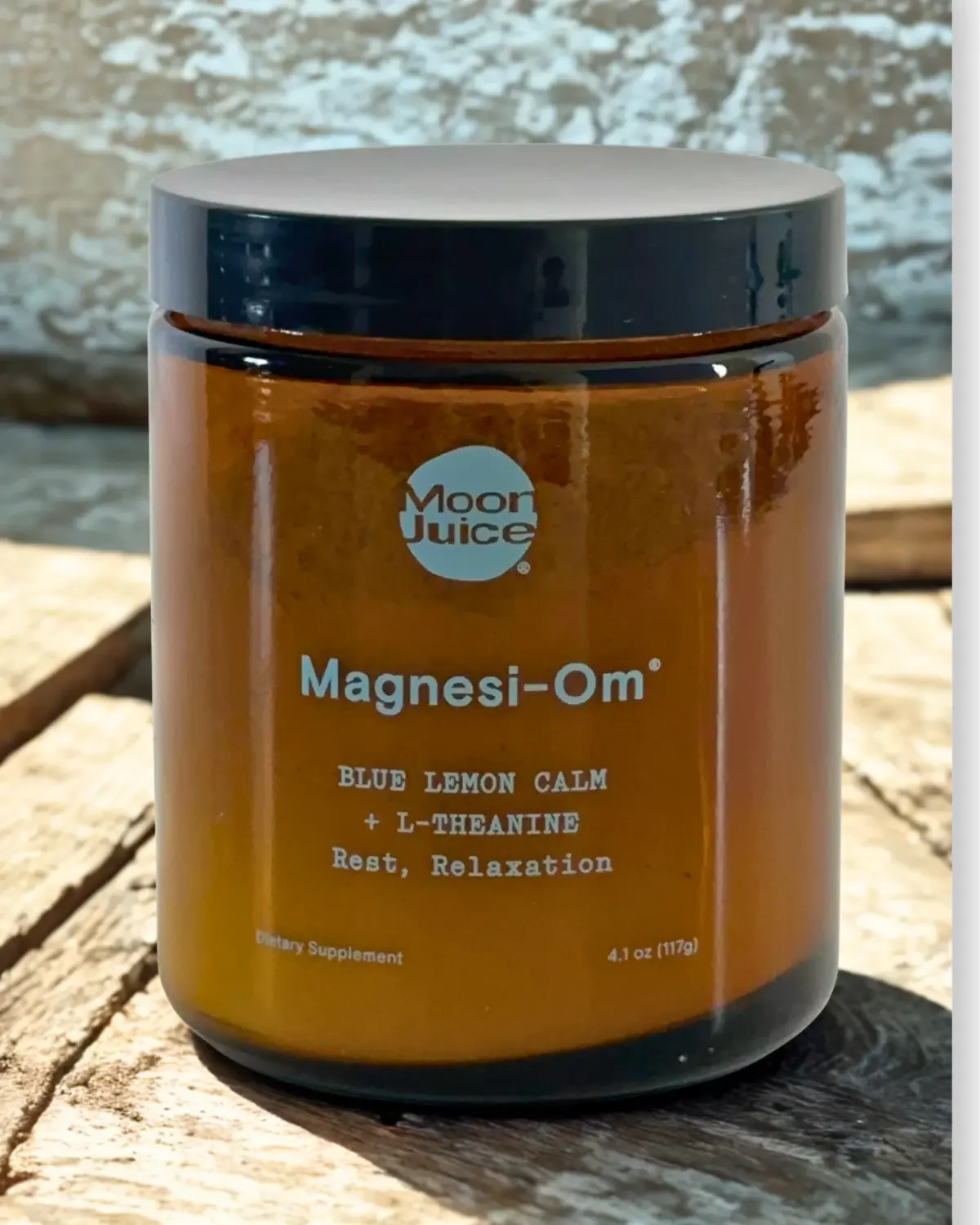 A brown jar labeled Moon Juice Magnesi-Om with ingredients Blue Lemon Calm and L-Theanine, for rest and relaxation, sitting on a wooden surface outdoors.